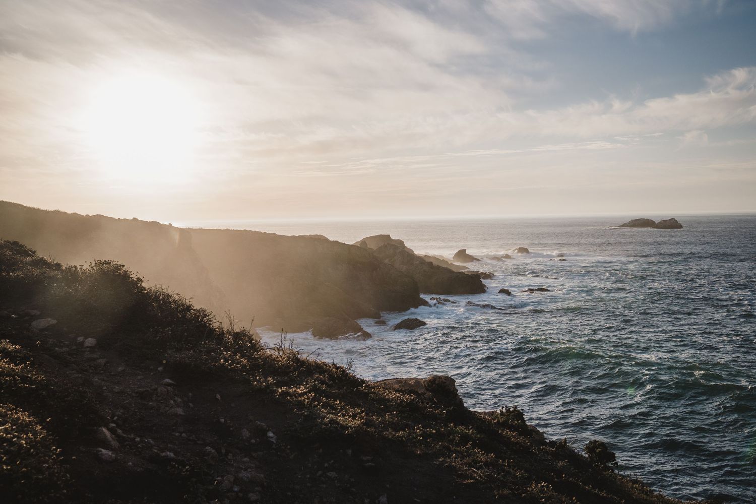 Dramatic coastal landscape at sunset with rocky cliffs overlooking a rugged Pacific Ocean shoreline and distant islands.
