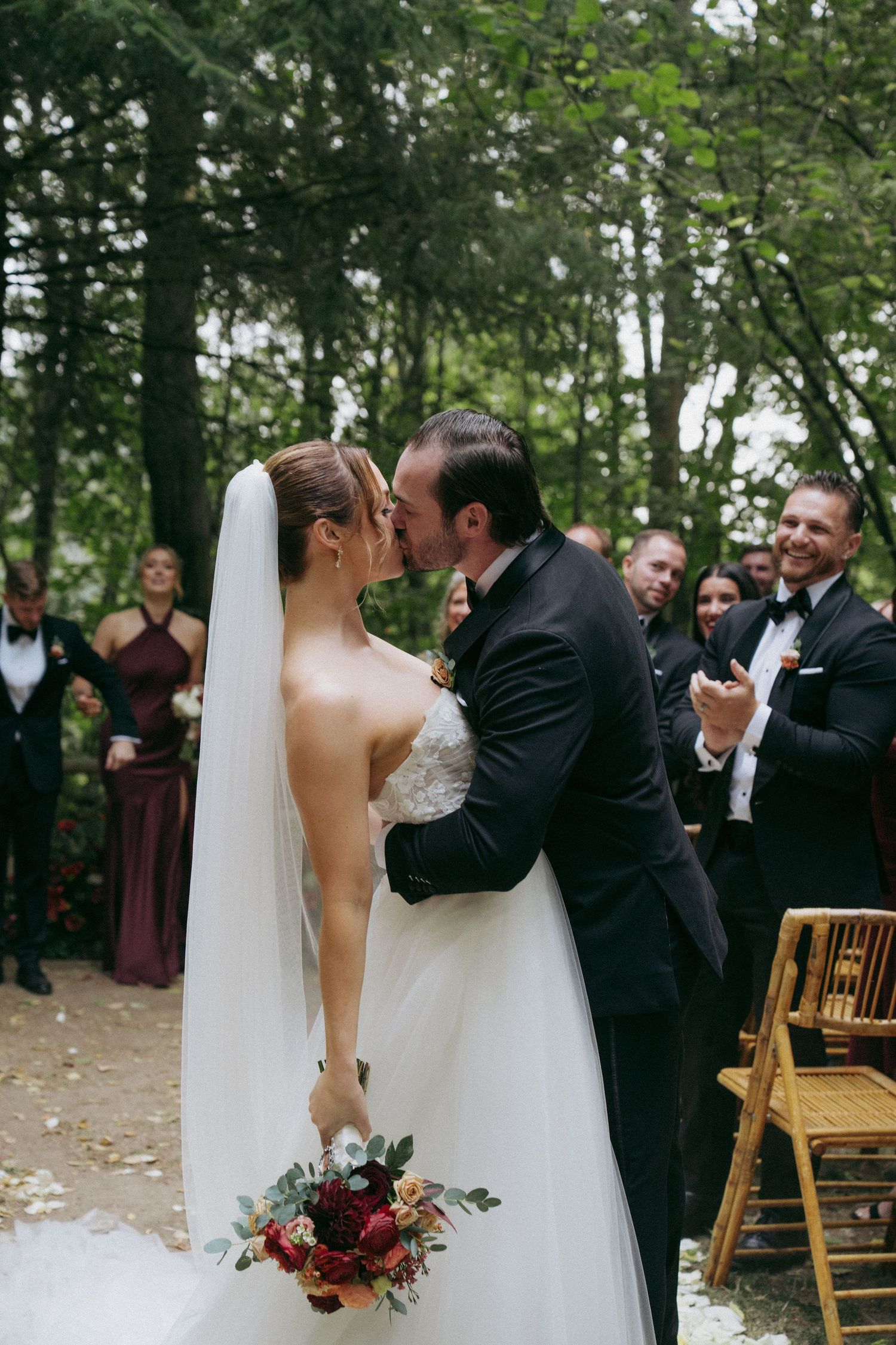 A romantic wedding ceremony kiss in a wooded outdoor setting with guests looking on.