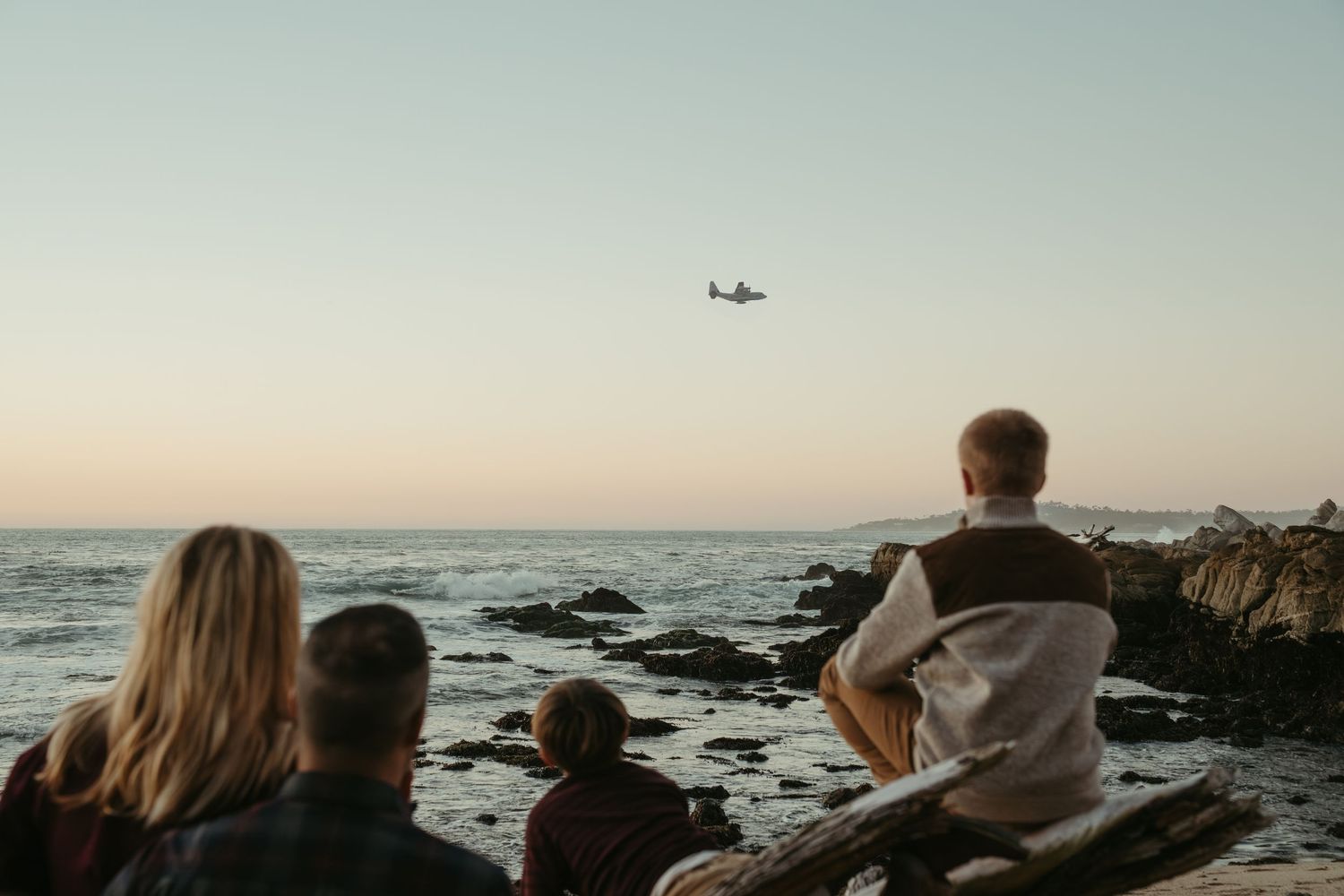 People watch a small airplane fly over the ocean from rocky coastal viewpoint at dusk.