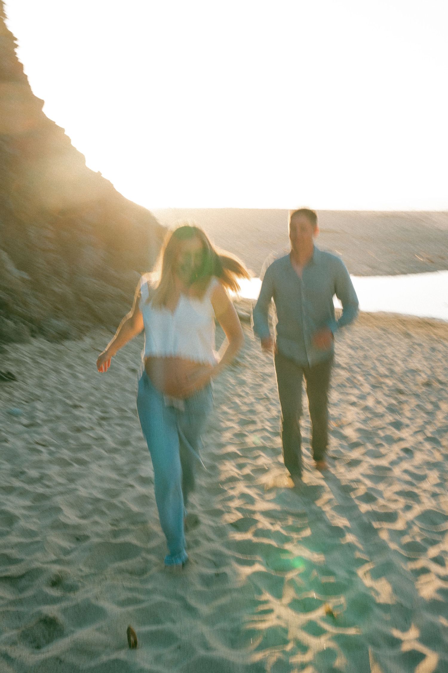 Couple running playfully on beach sand with golden sunlight filtering through rocks in background.