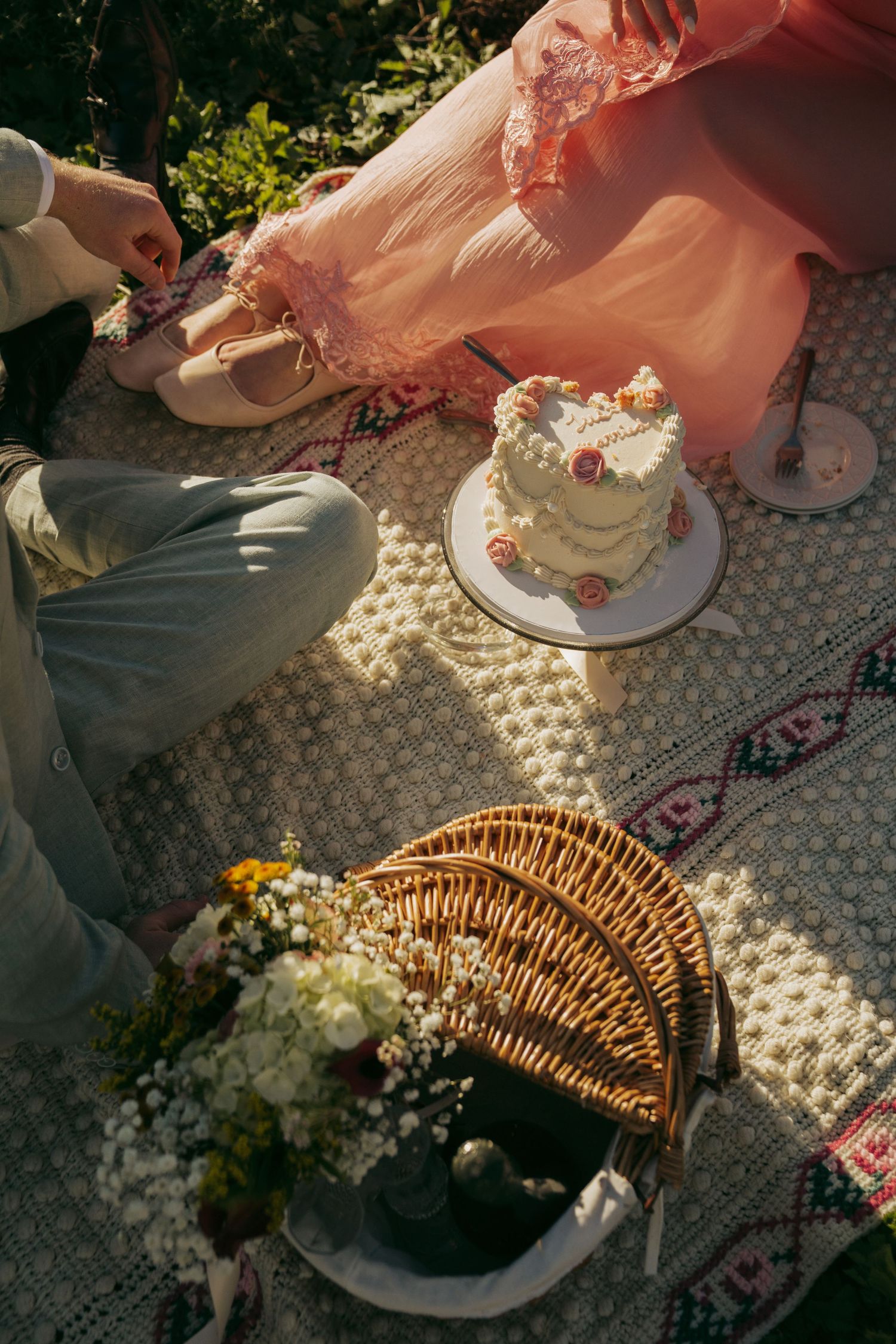 Aerial view of a romantic picnic with a decorated cake, wicker basket, flowers, and two people on a patterned blanket.