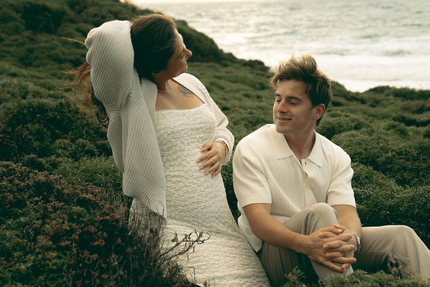 A couple sits together on a lush green hillside near the ocean, sharing a tender moment in white outfits.