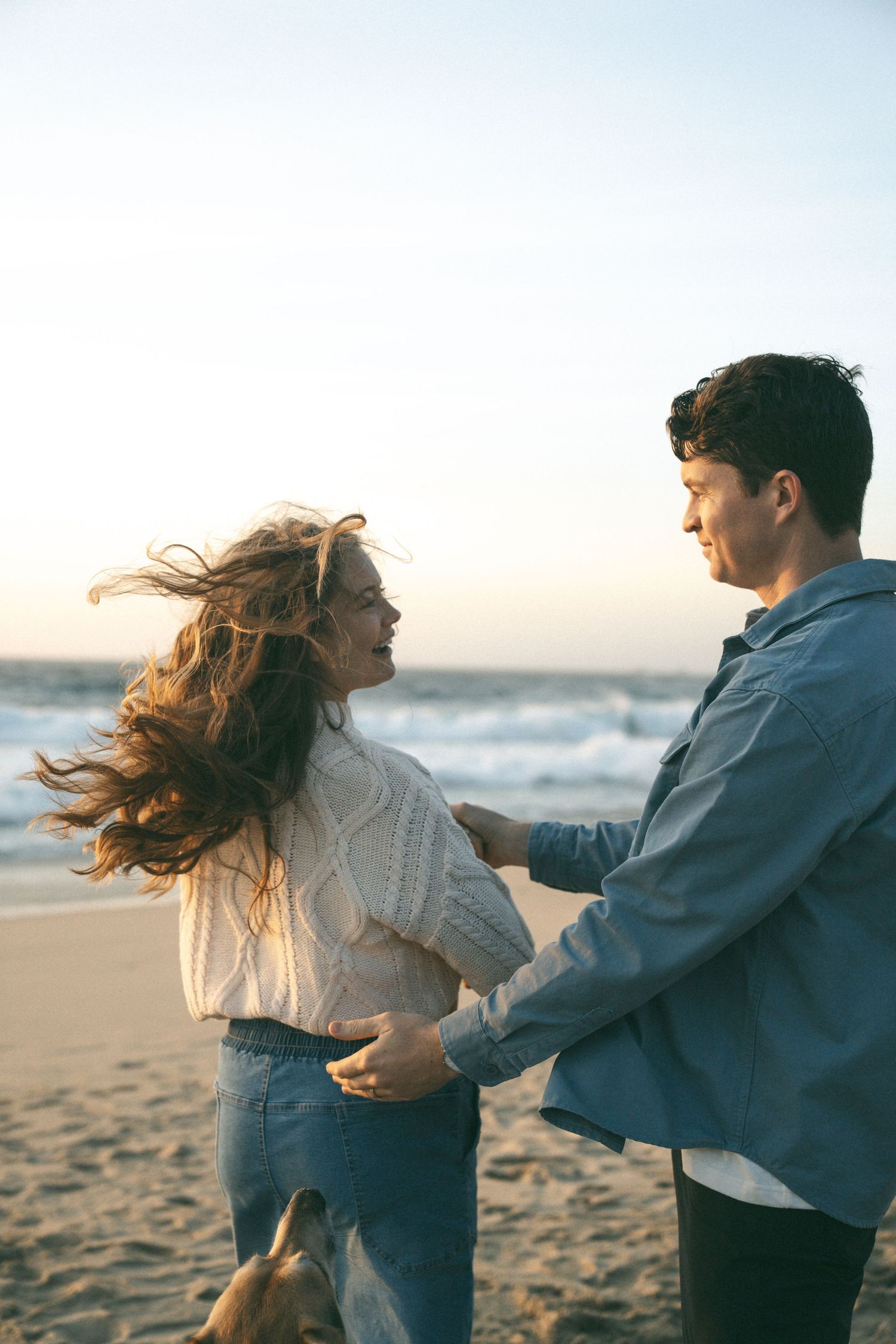 Couple holding hands and dancing on a windy beach at sunset, woman in white sweater and jeans smiling.