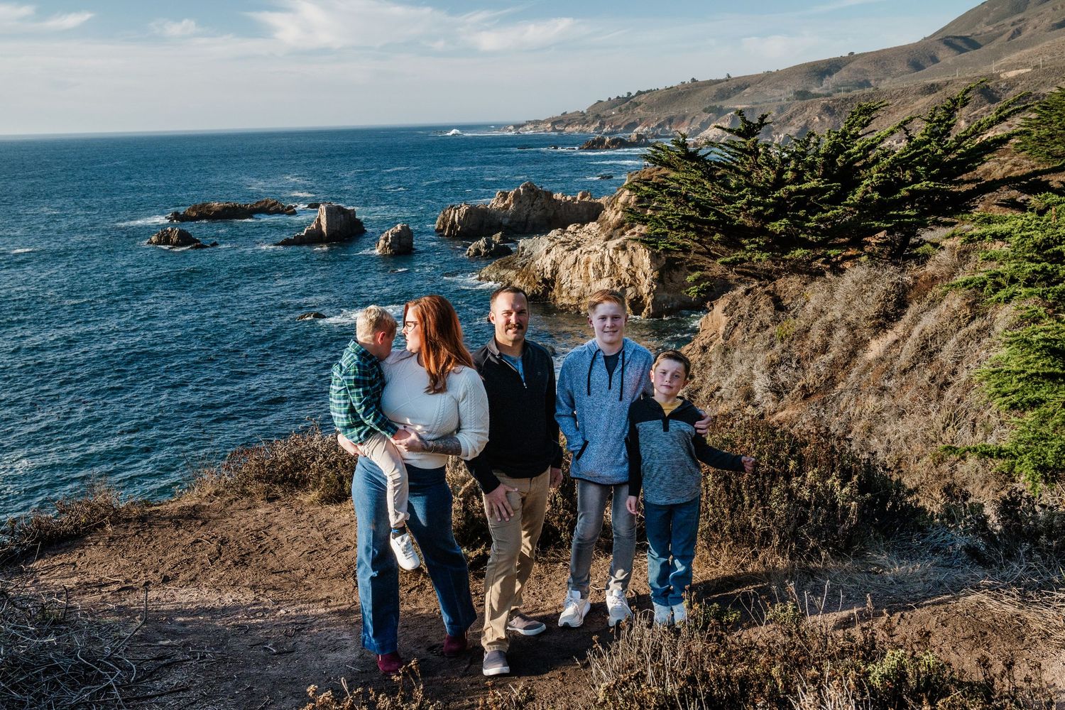 Family enjoying scenic coastal views along rugged Big Sur cliffs with Pacific Ocean and rocky coastline in background.