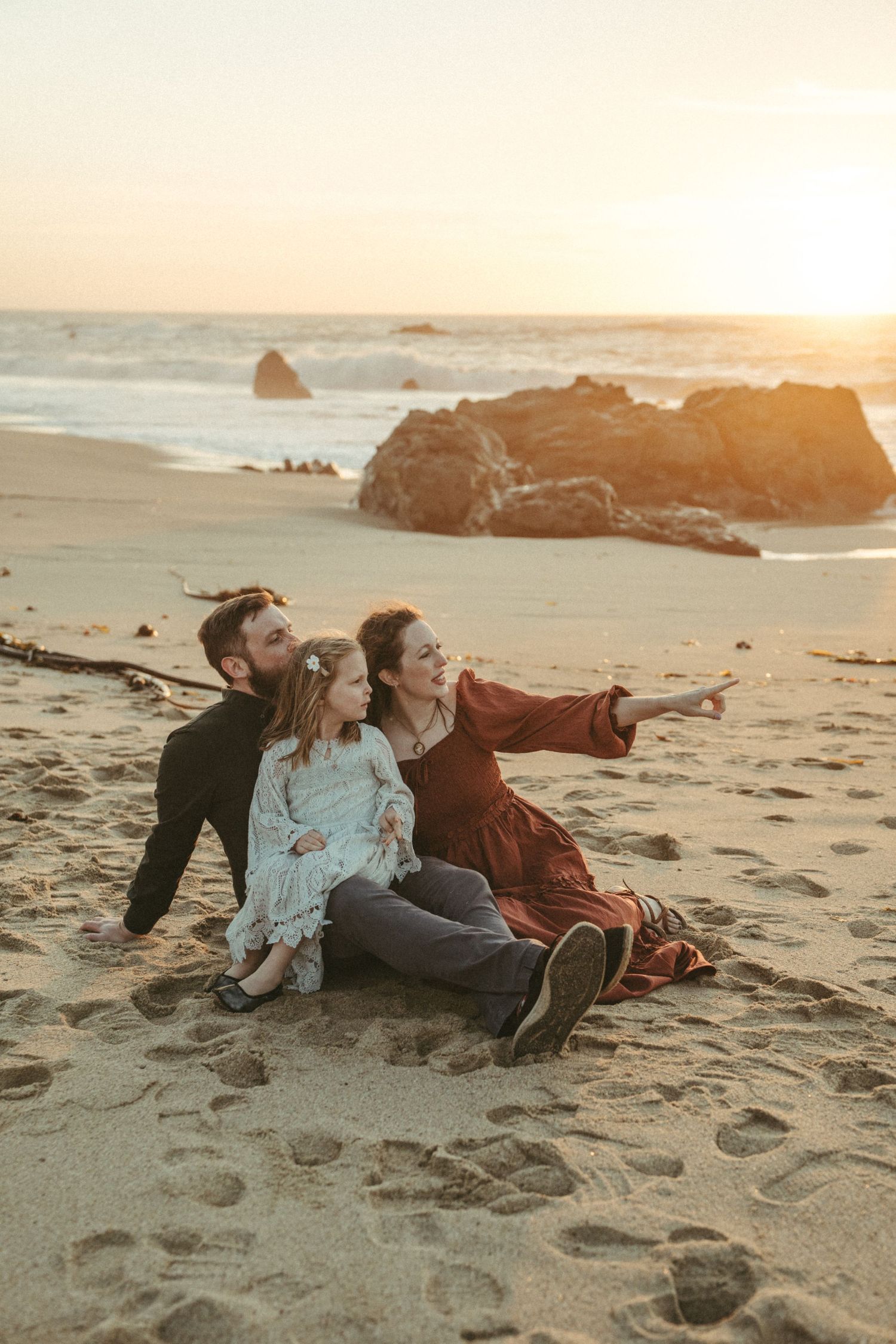 A couple relaxes together on a sandy beach at sunset with ocean waves and rocks in the background.