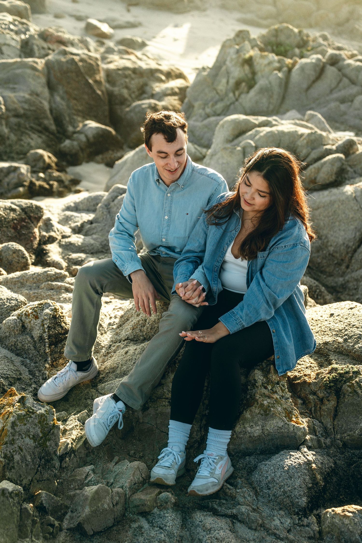 Couple in denim sitting together on rocky coastal outcrop at sunset.
