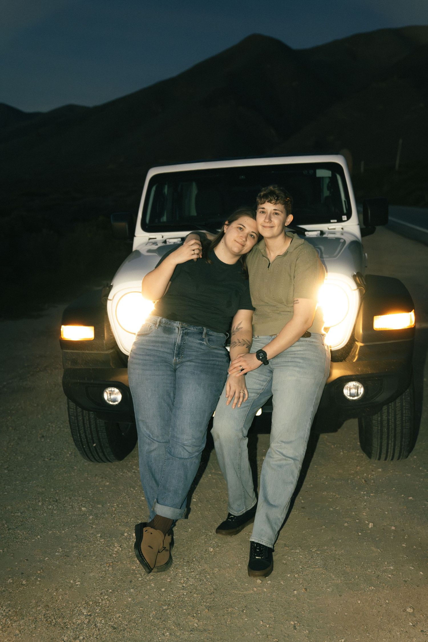 A couple poses together against their white Jeep at night with mountains silhouetted in the background.