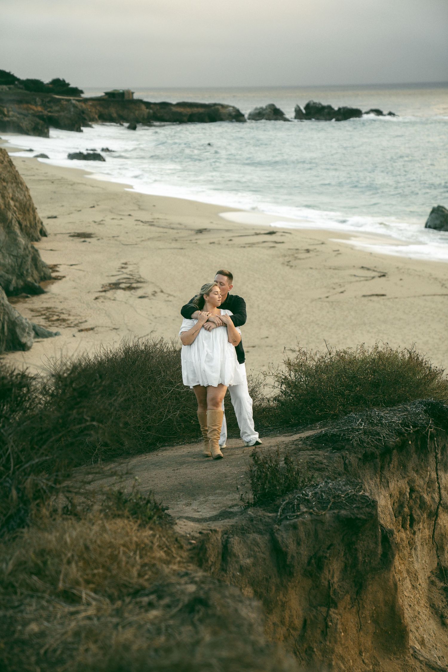 A couple embraces on a scenic coastal path overlooking a rocky beach with crashing waves.