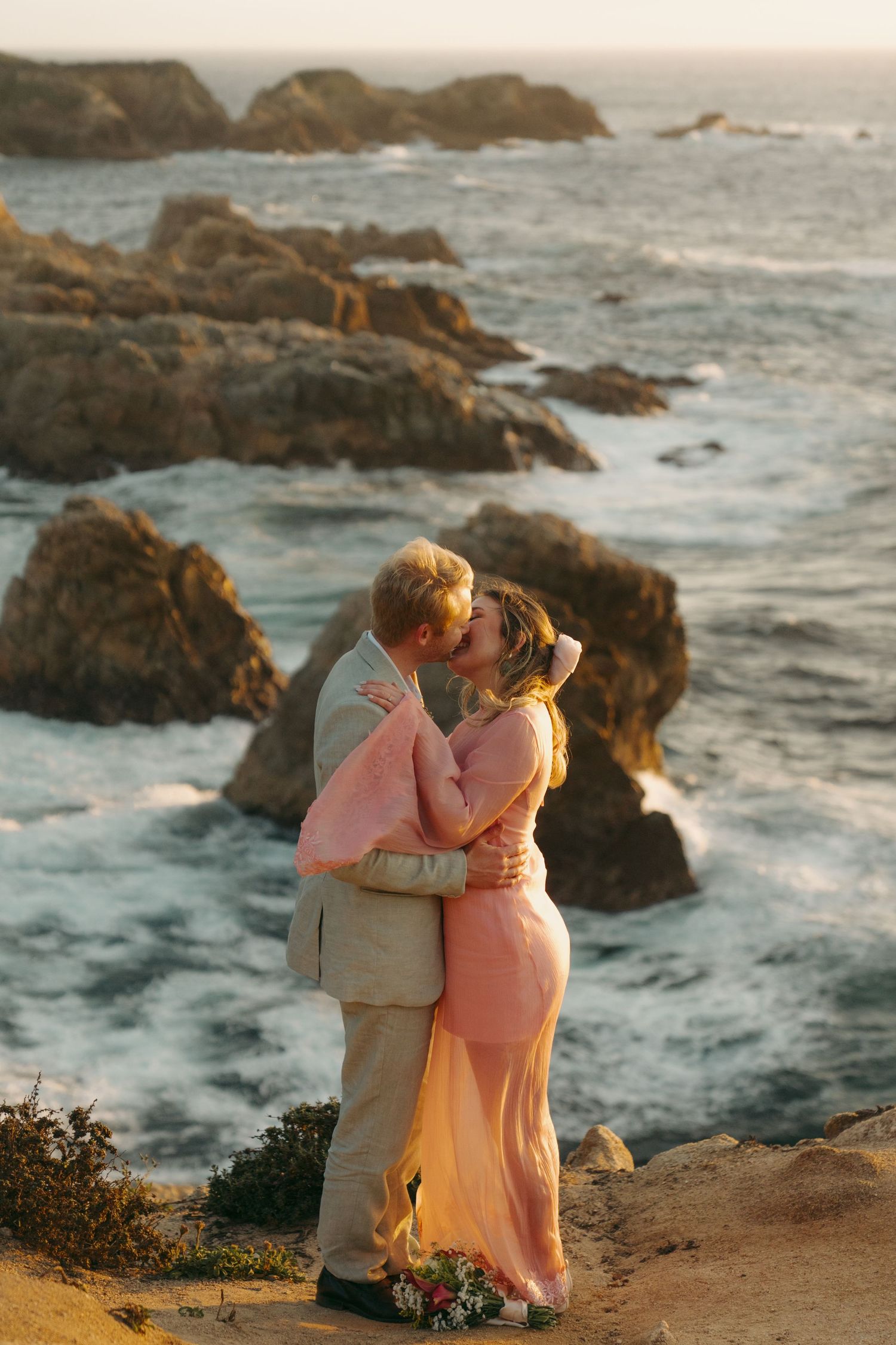 Couple sharing a romantic kiss on a rocky coastal cliff at sunset, dressed in elegant pink and beige wedding attire.