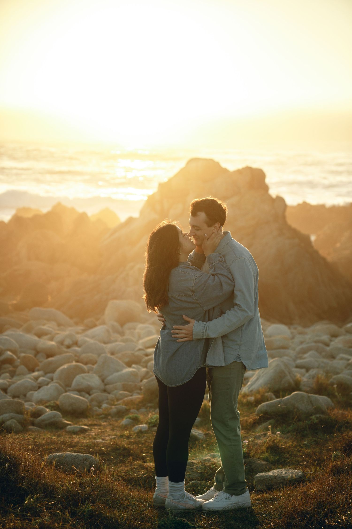 Couple embracing on rocky coastal terrain during golden sunset.