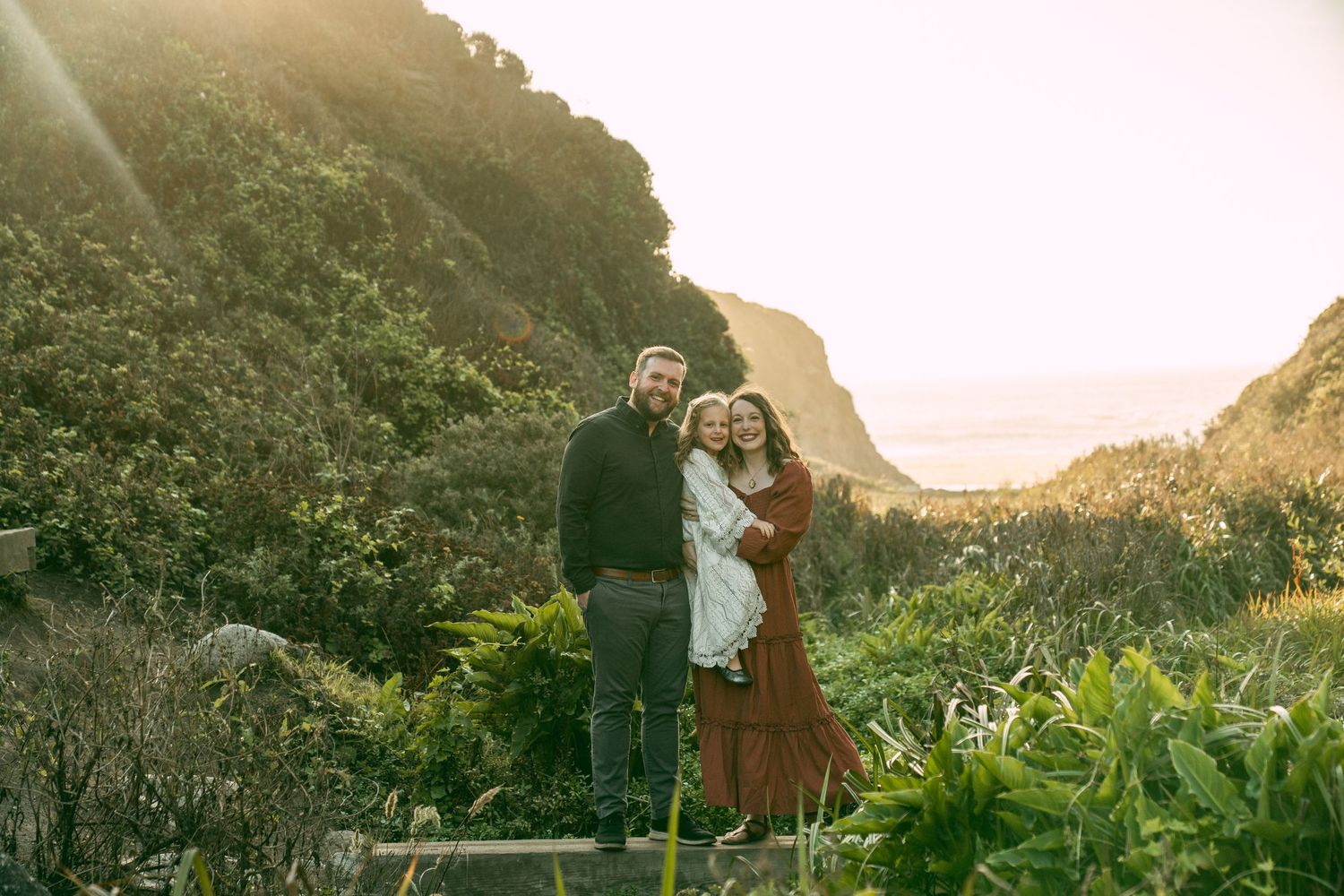 A couple stands together against a backdrop of dramatic coastal cliffs and lush vegetation at golden hour.