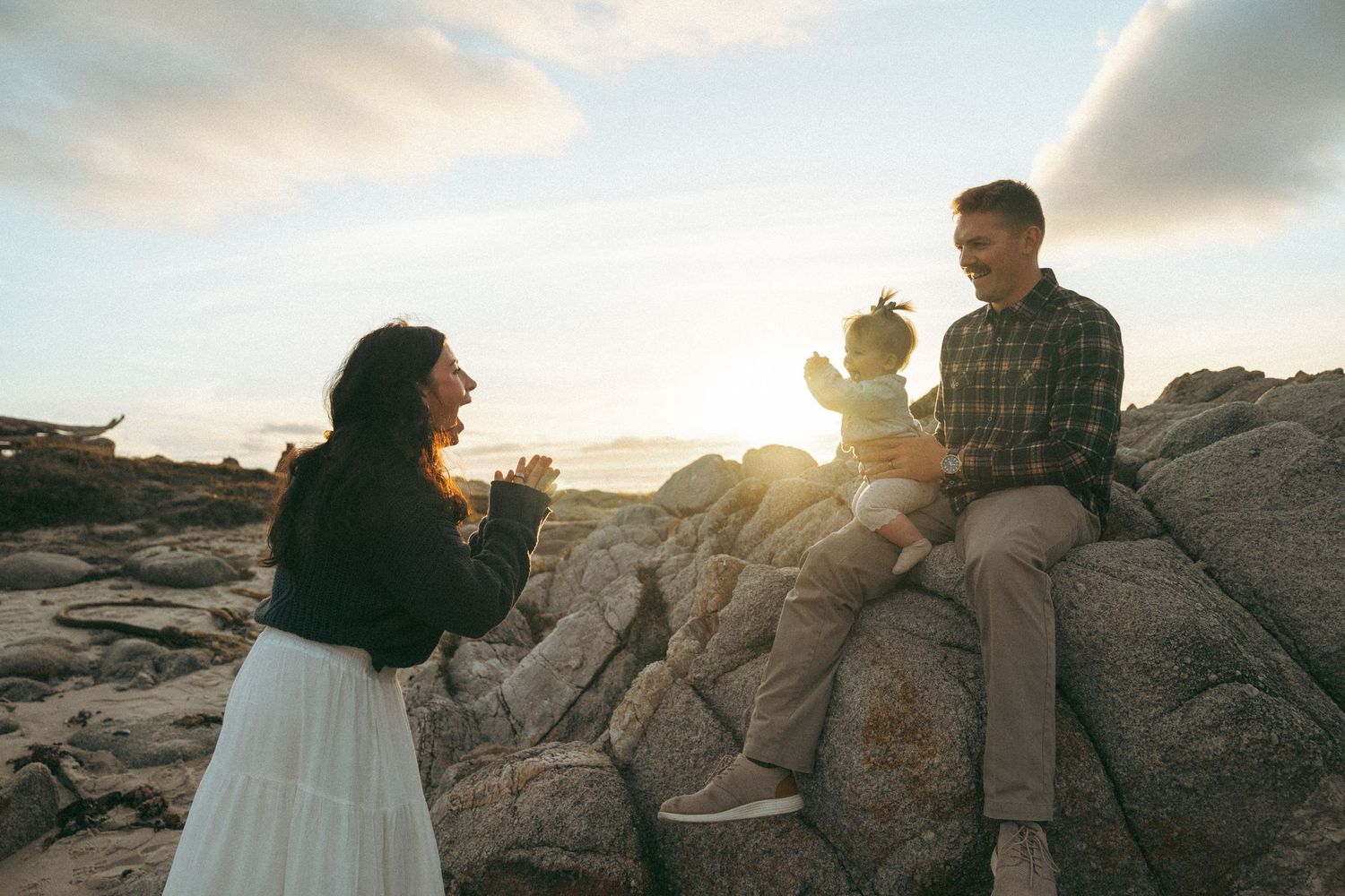 Couple enjoying sunset together on rocky coastal landscape as sunlight streams through dramatic clouds.
