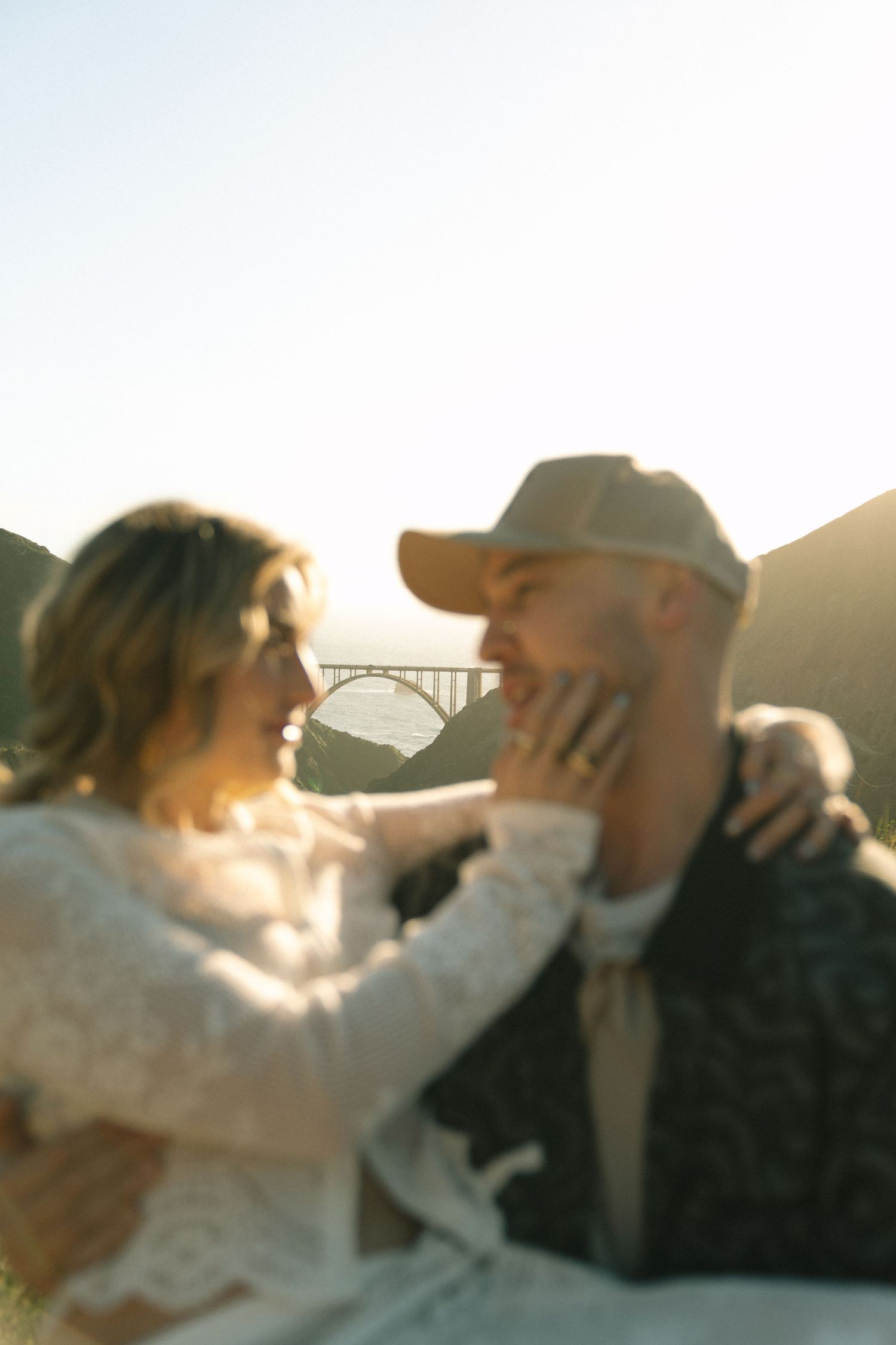 Couple sharing a tender moment outdoors with Bixby Bridge in the background at sunset.