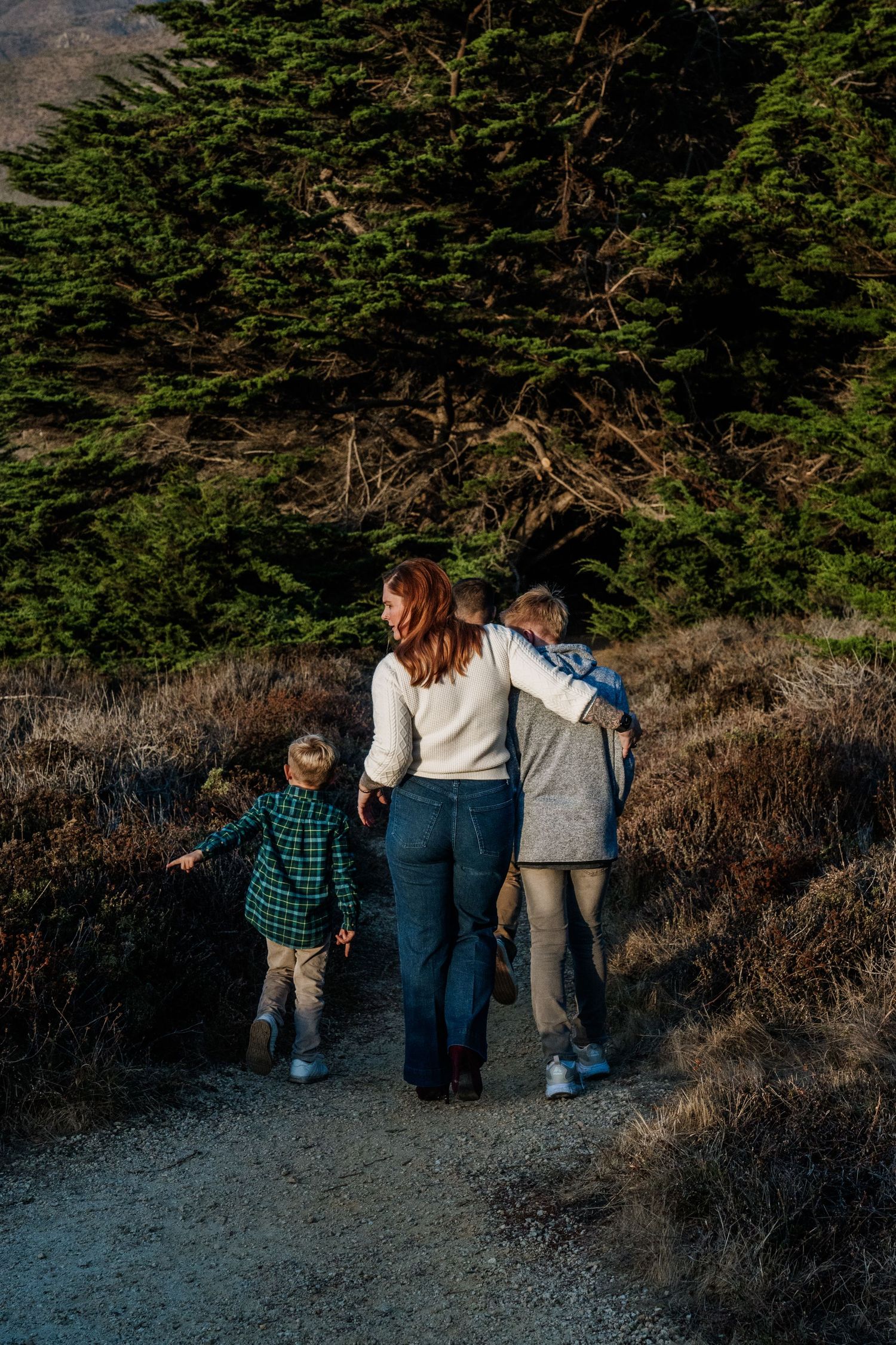 Three people walking together on a scenic trail surrounded by evergreen trees and coastal brush at sunset.