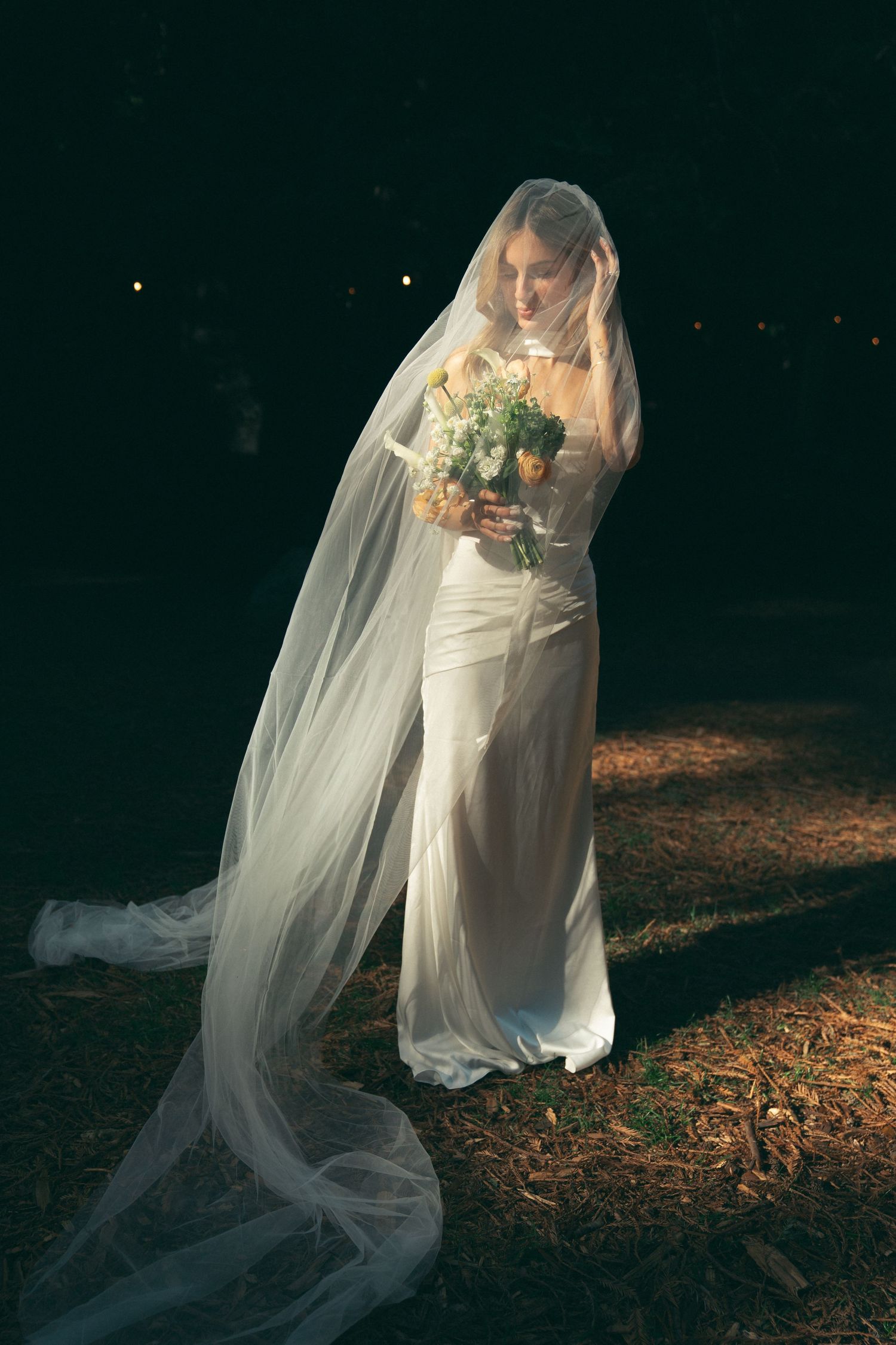 A bride in a white wedding dress and long flowing veil holds a bouquet while standing in dramatic evening lighting.