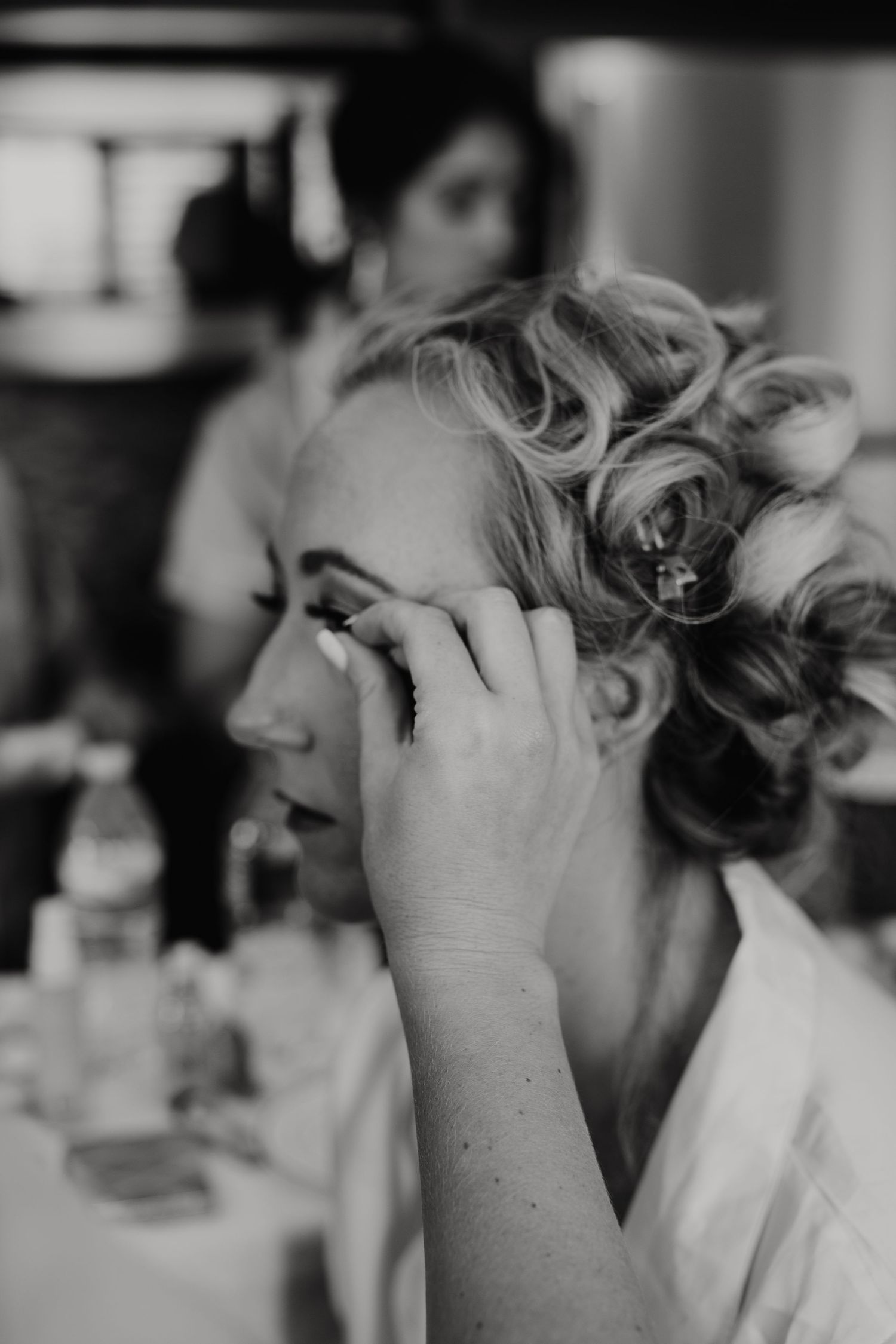 Black and white photo of a woman getting her makeup done, wearing a white robe while sitting at a dressing table.