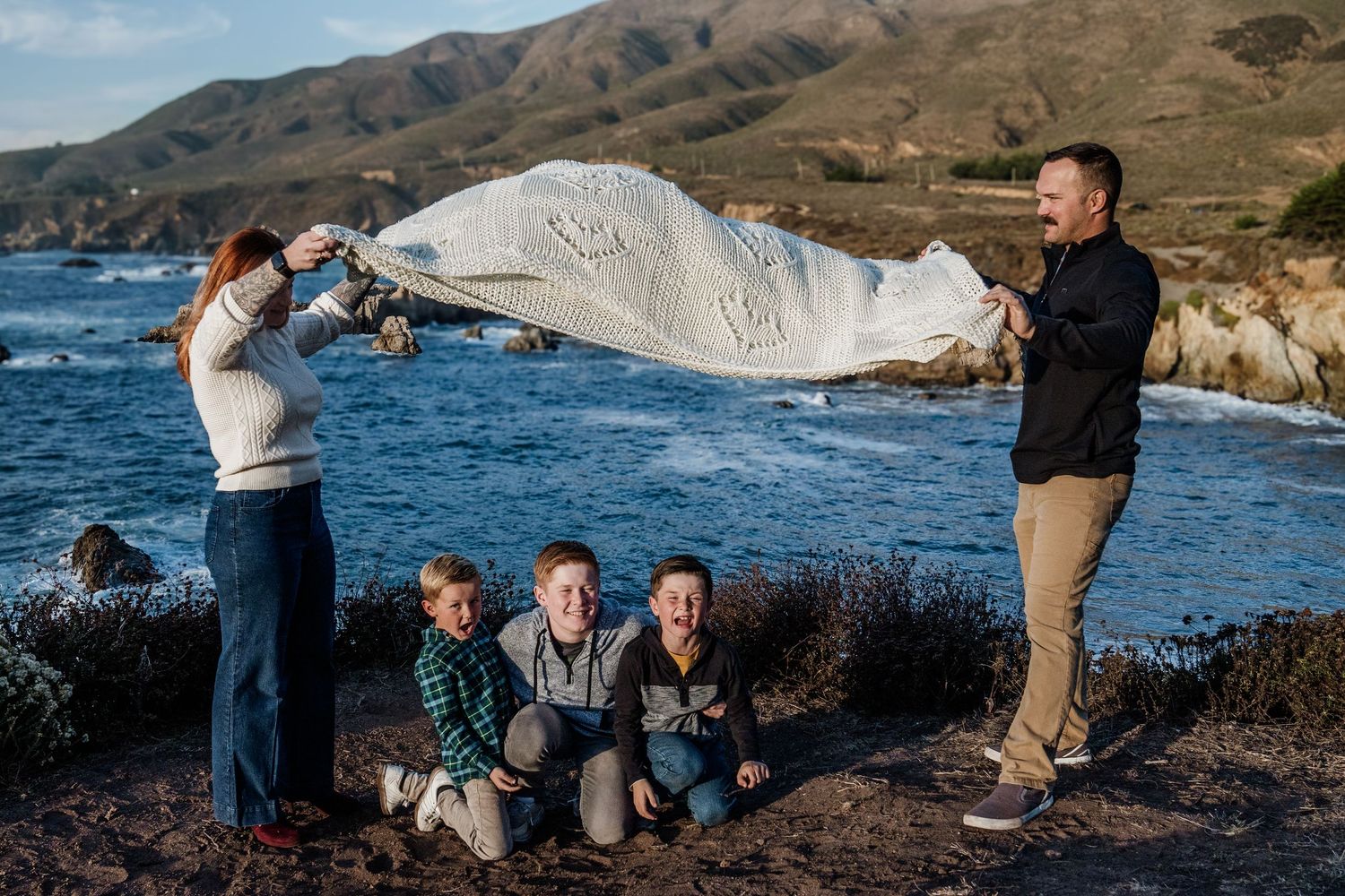 A family plays with a white blanket along rocky coastal cliffs with blue ocean waves and mountains in the background.