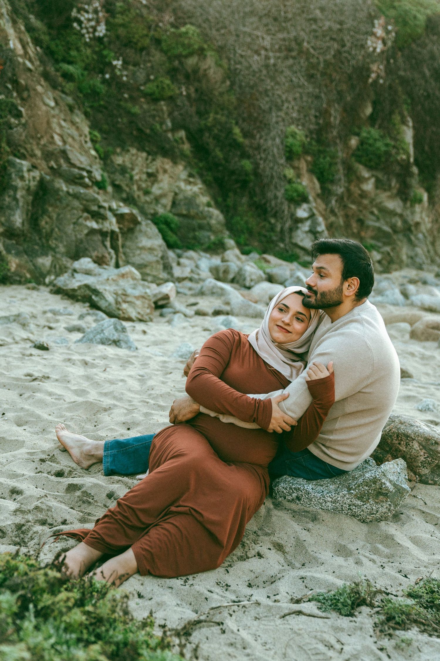 A couple relaxes together on a sandy beach against rocky cliffs.