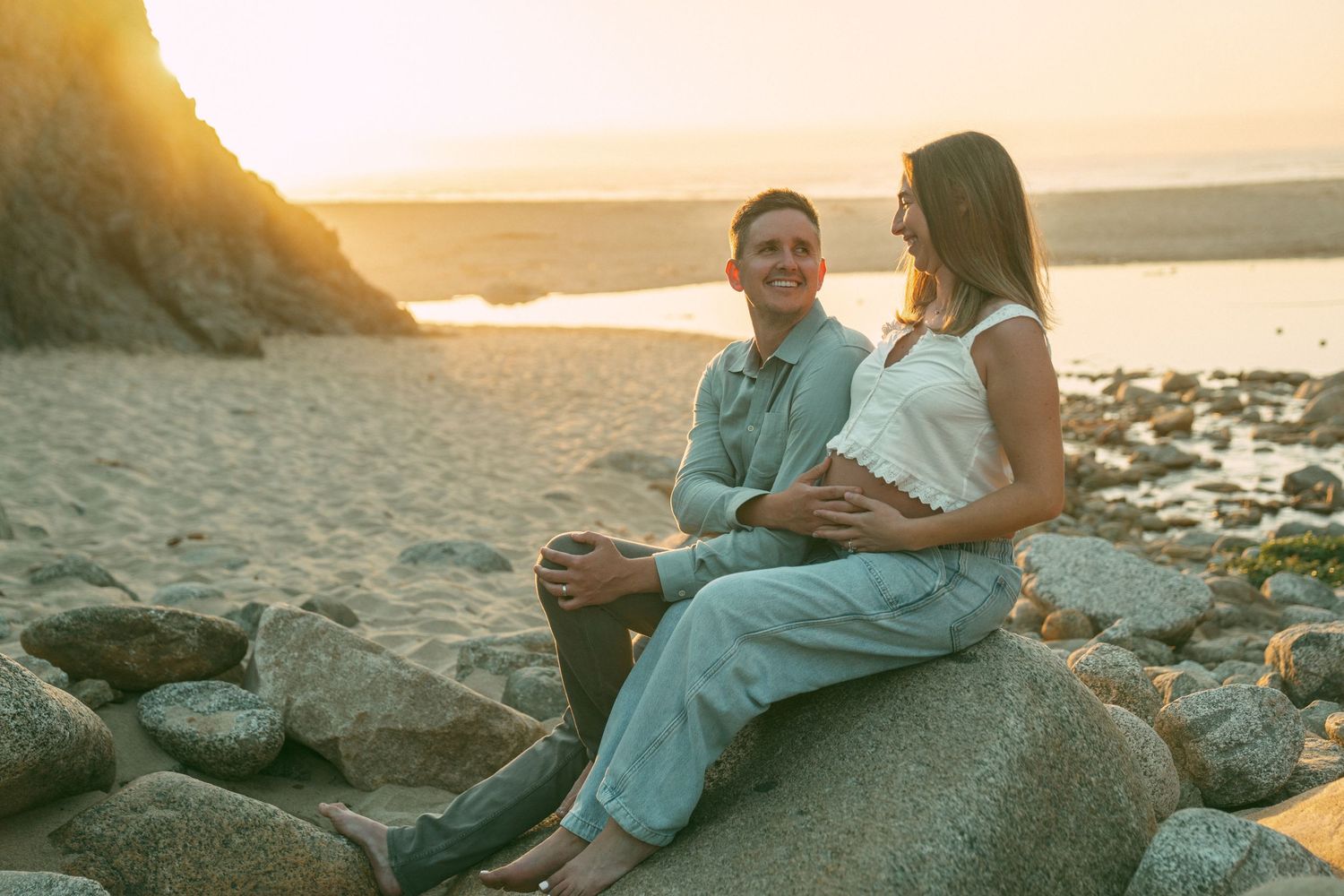 A couple sits together on coastal rocks during golden hour sunset by the ocean.