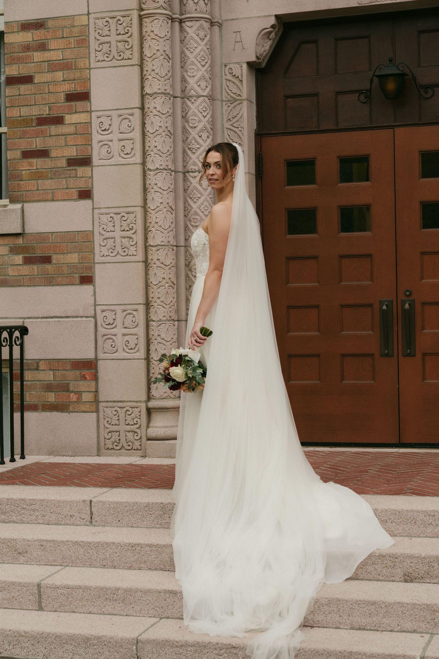 An elegant bride in a flowing white gown stands on stone steps beside ornate architectural columns and wooden doors.