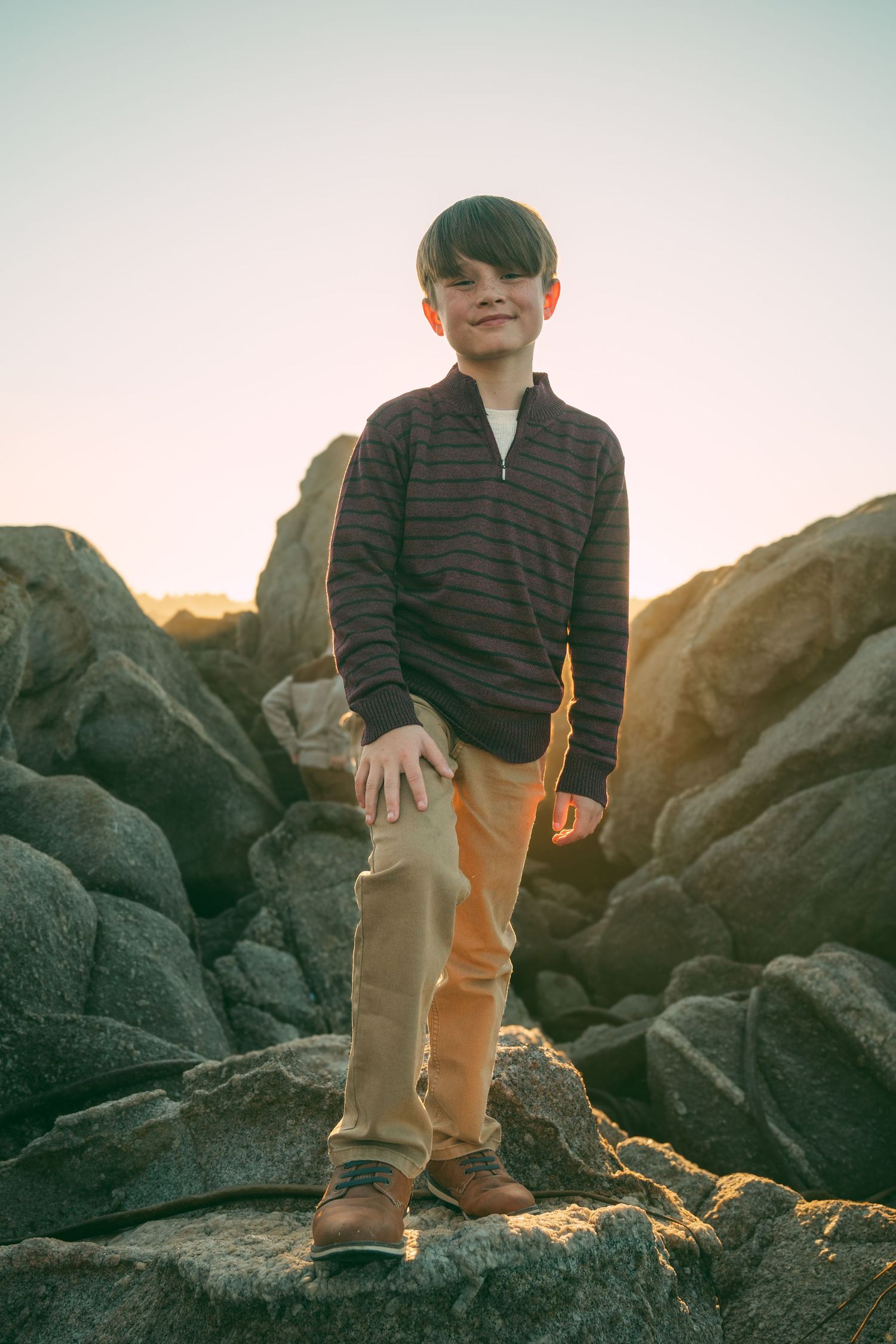 Person wearing casual clothing stands on rocky coastal terrain at sunset with warm golden light illuminating the scene.