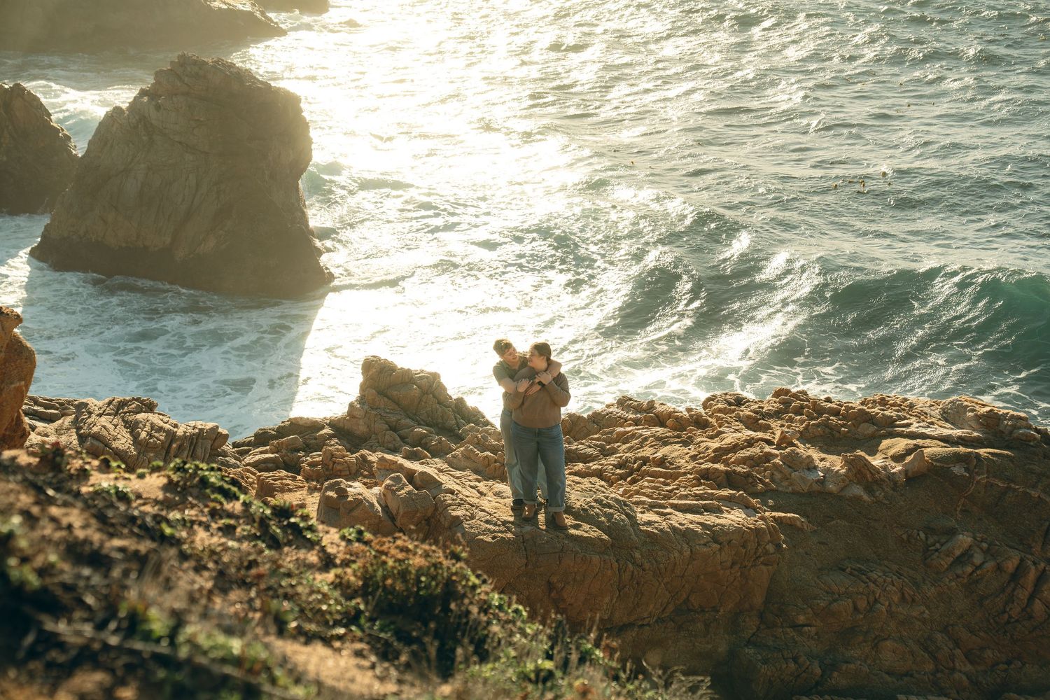 Rocky coastline with waves crashing against cliffs during sunset with figures in distance.