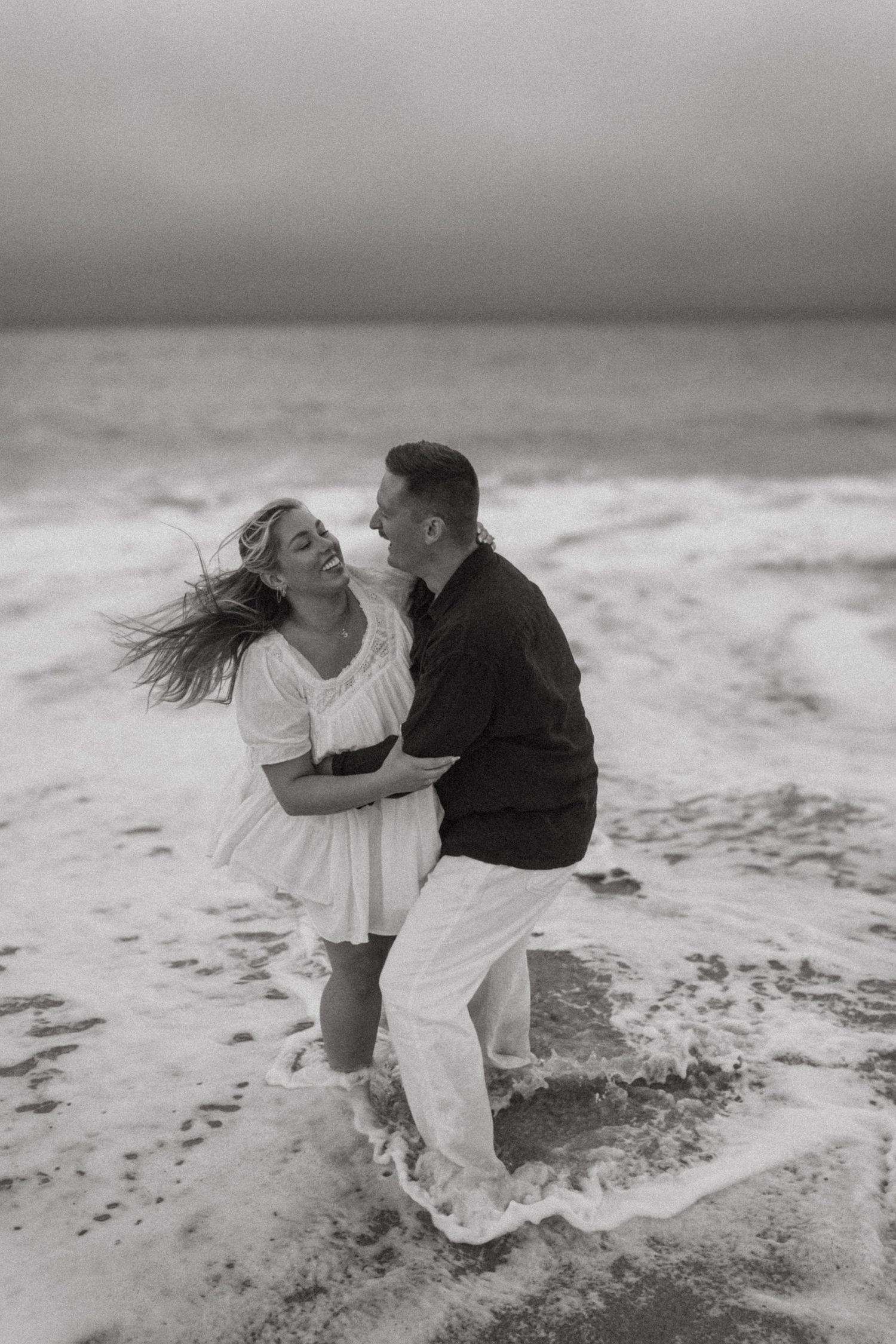 A couple shares an intimate moment on a cloudy beach with waves crashing in the background.