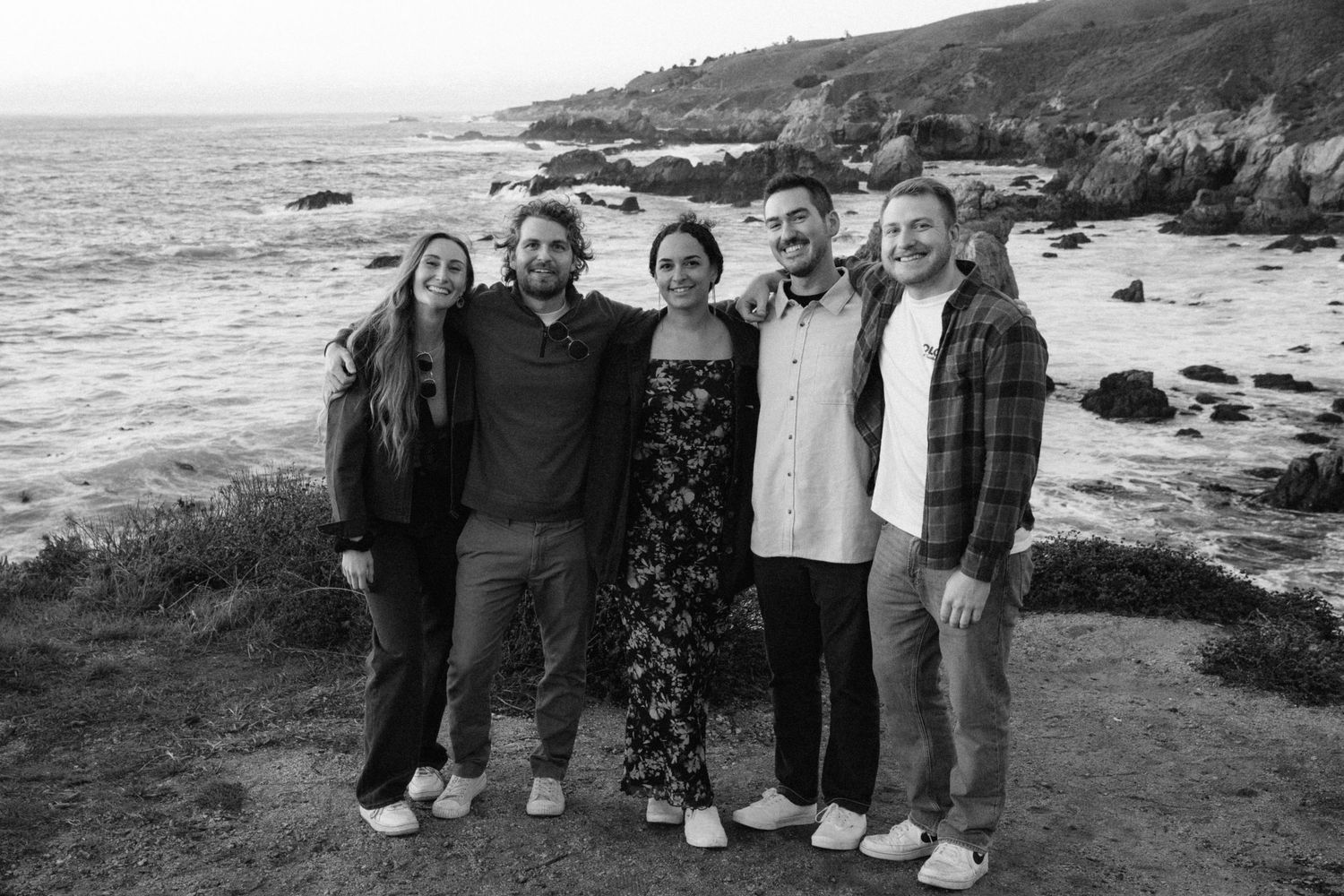 A group gathers on a scenic coastal path overlooking rocky cliffs and ocean waves during golden hour.