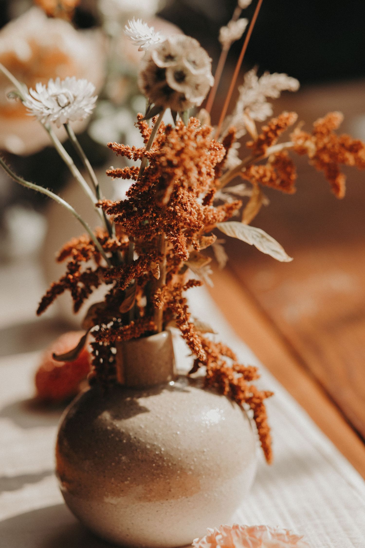 Small white ceramic bud vase holds delicate dried orange and white flowers on a rustic wooden table.