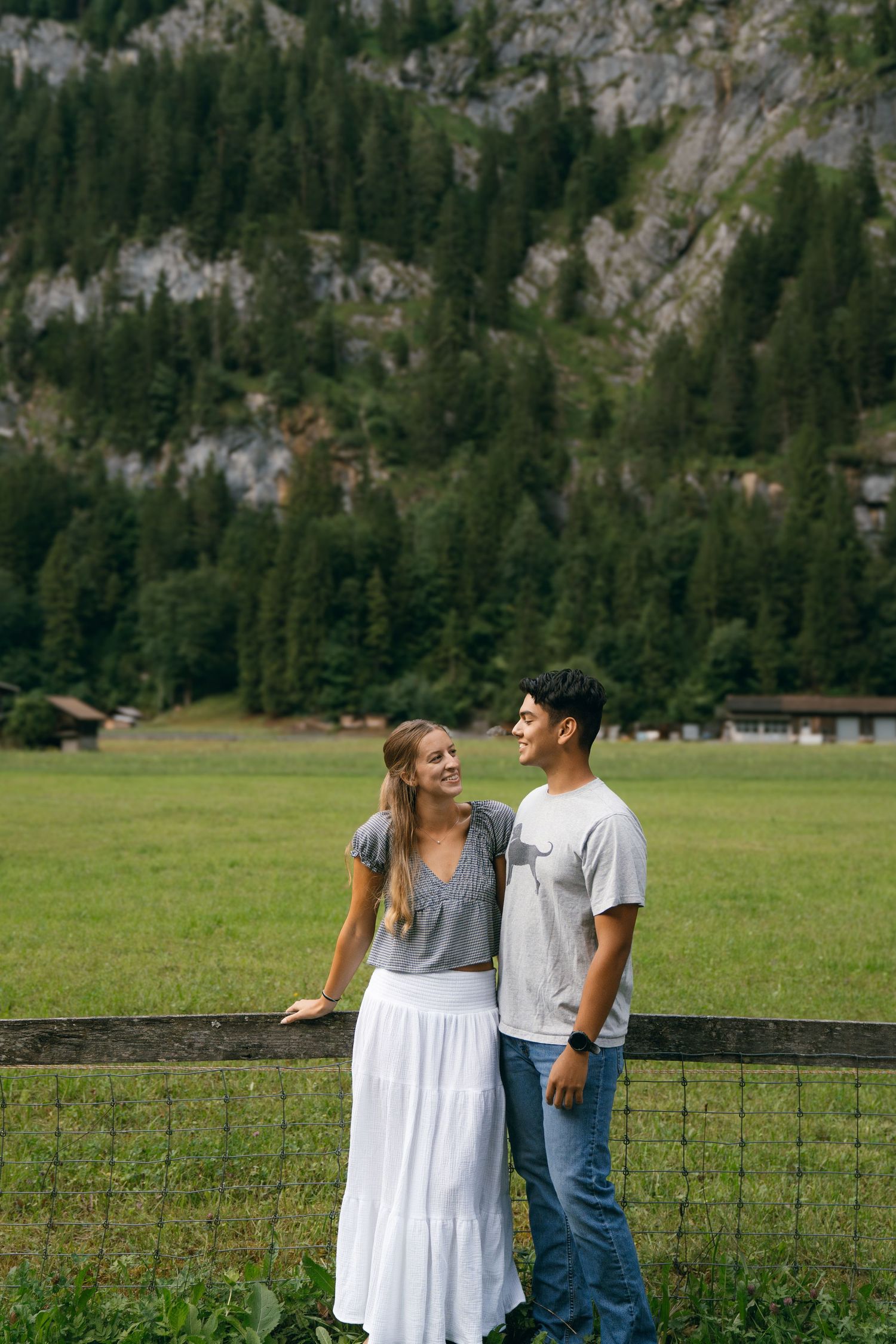 A couple shares a joyful moment together while standing by a fence in a scenic mountain valley with evergreen forests.