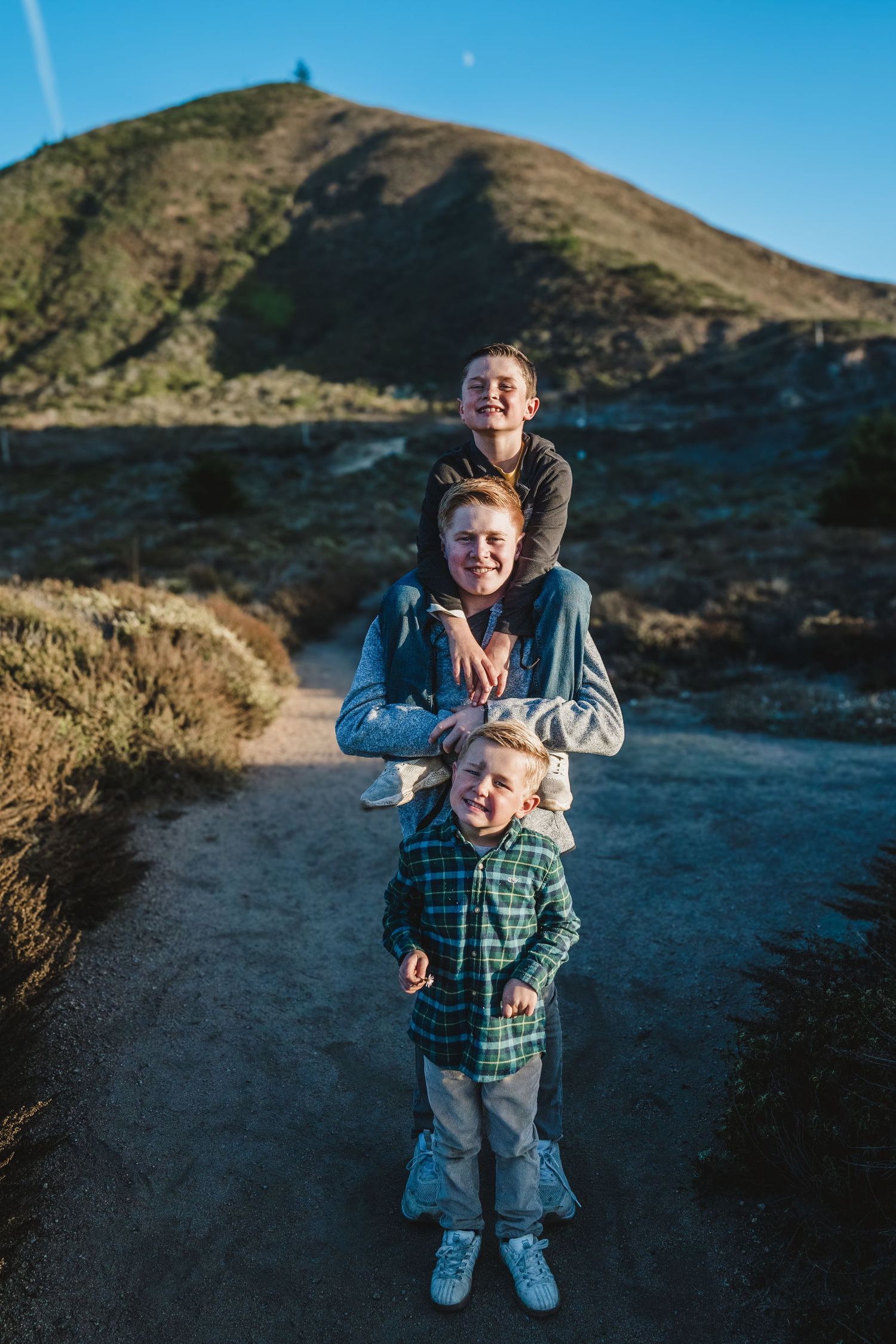 Three people pose playfully on a hiking trail with mountains and blue sky in the background.