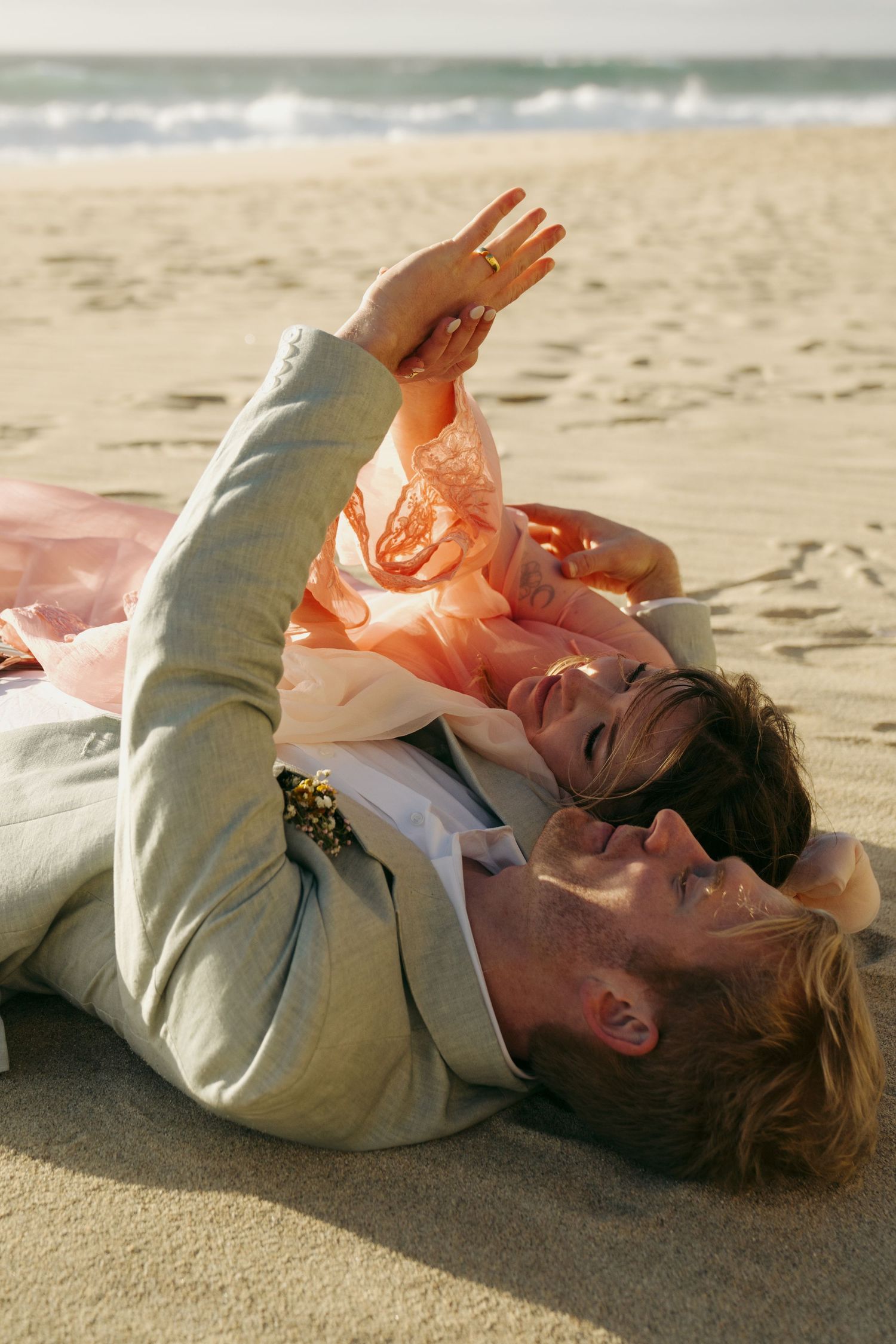 Couple in wedding attire lying on sandy beach, woman in pink dress resting on man in gray suit, ocean waves in background.