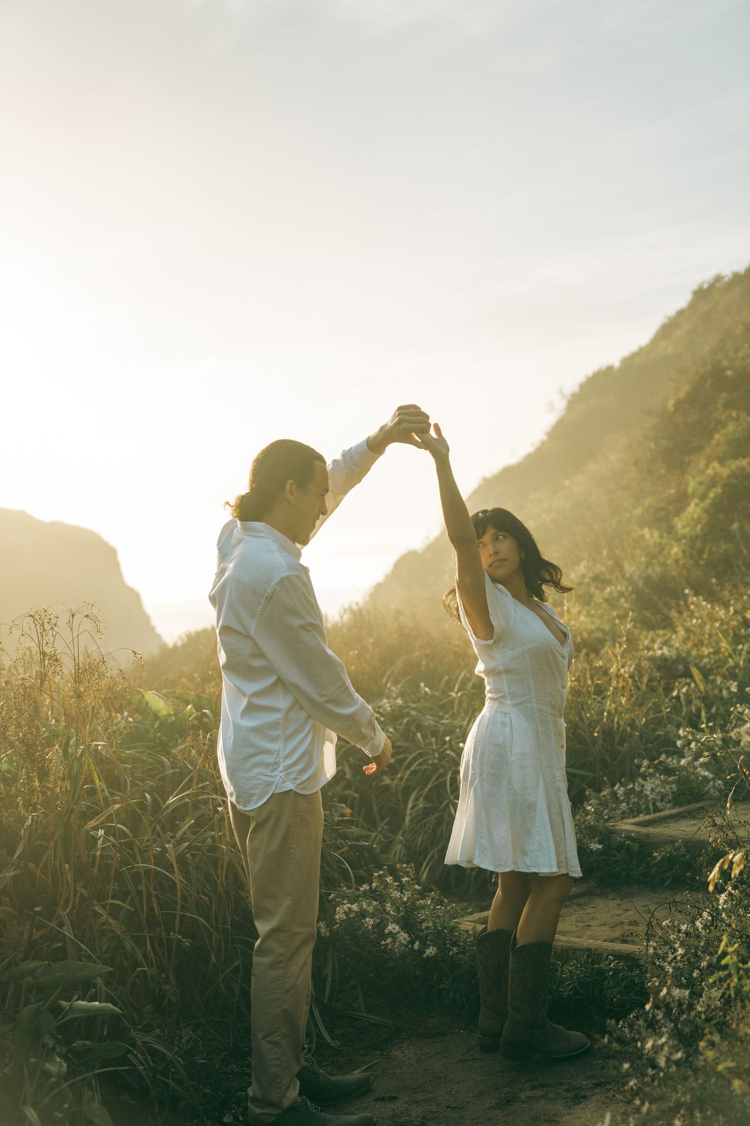 A couple in white clothing dances together in a sunlit meadow against a mountainous backdrop.
