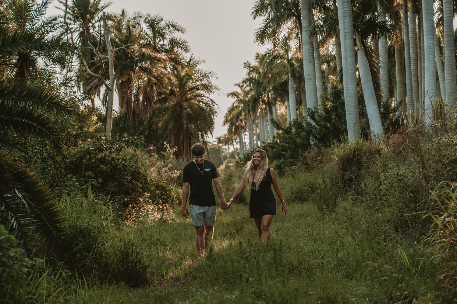 Two dark figures walk together on a lush path through a tropical palm tree grove at sunset.