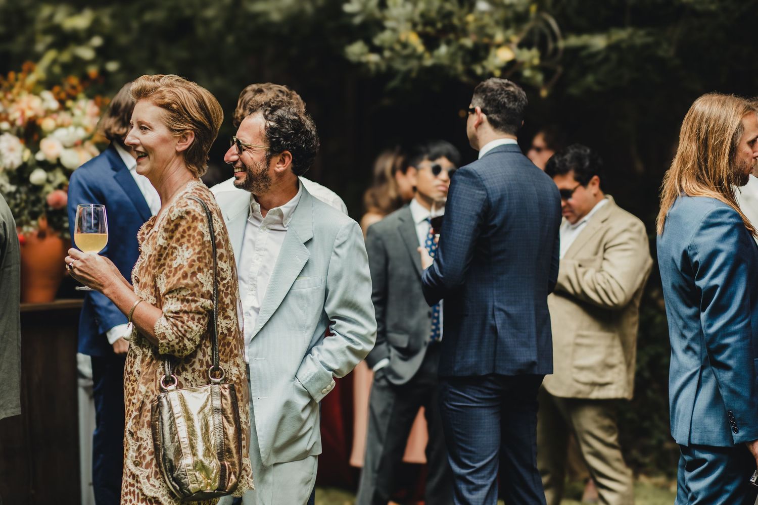 A group of wedding guests in formal attire stand and chat during an outdoor reception.
