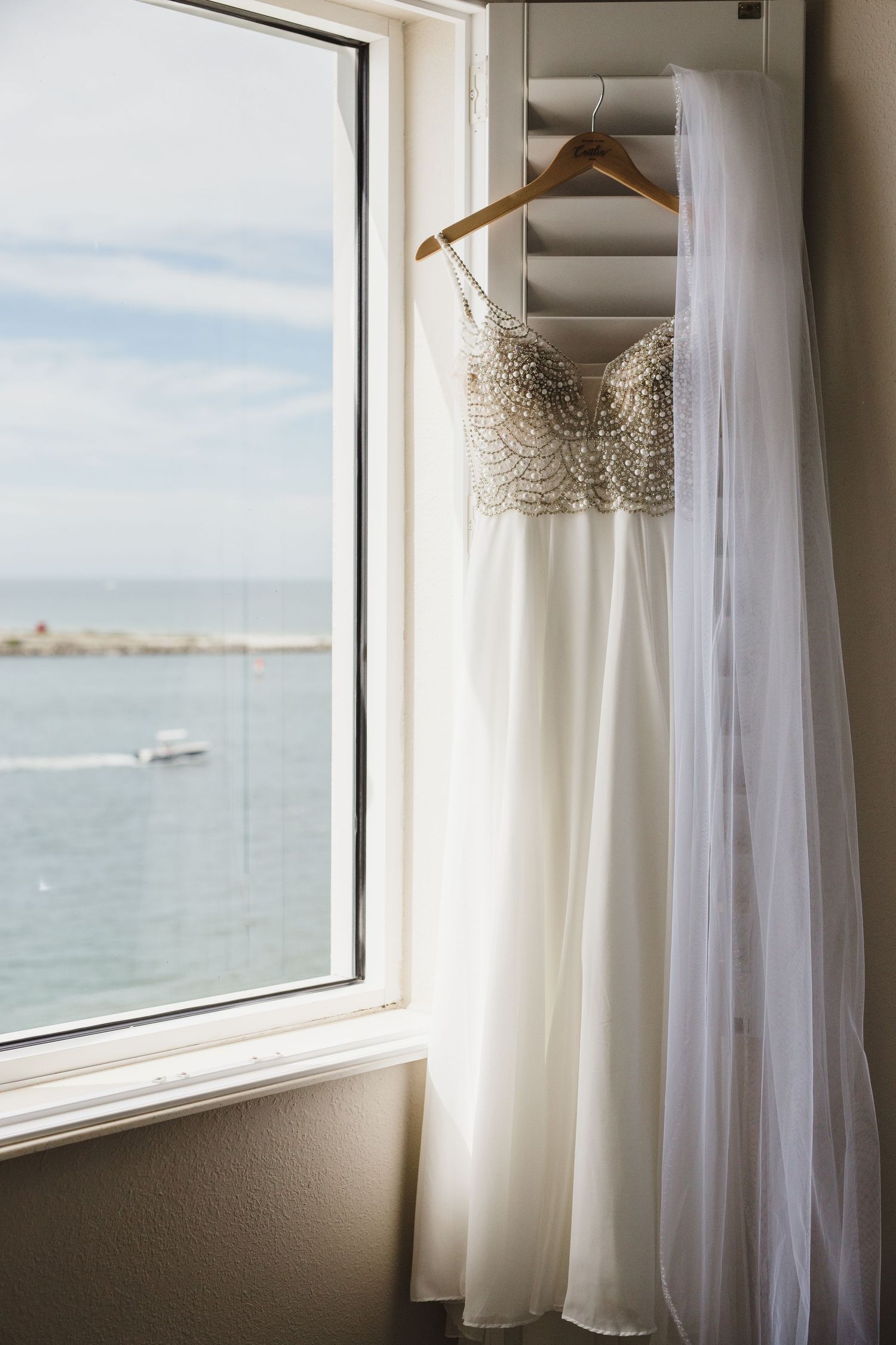 A white wedding dress hangs by a window overlooking a scenic ocean view on a bright sunny day.