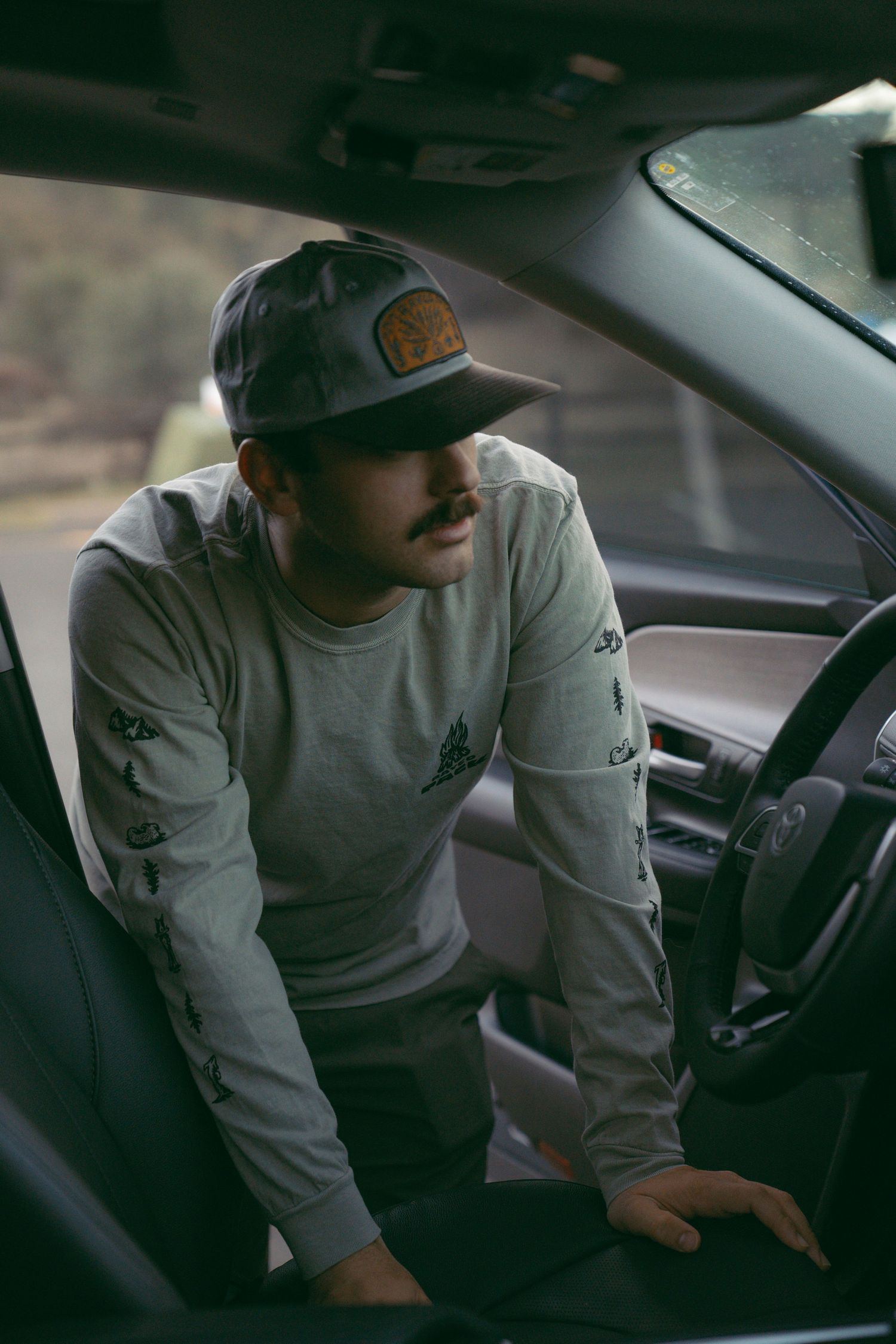 Person wearing a gray sweatshirt and baseball cap sits in the driver's seat of a car looking to the side.