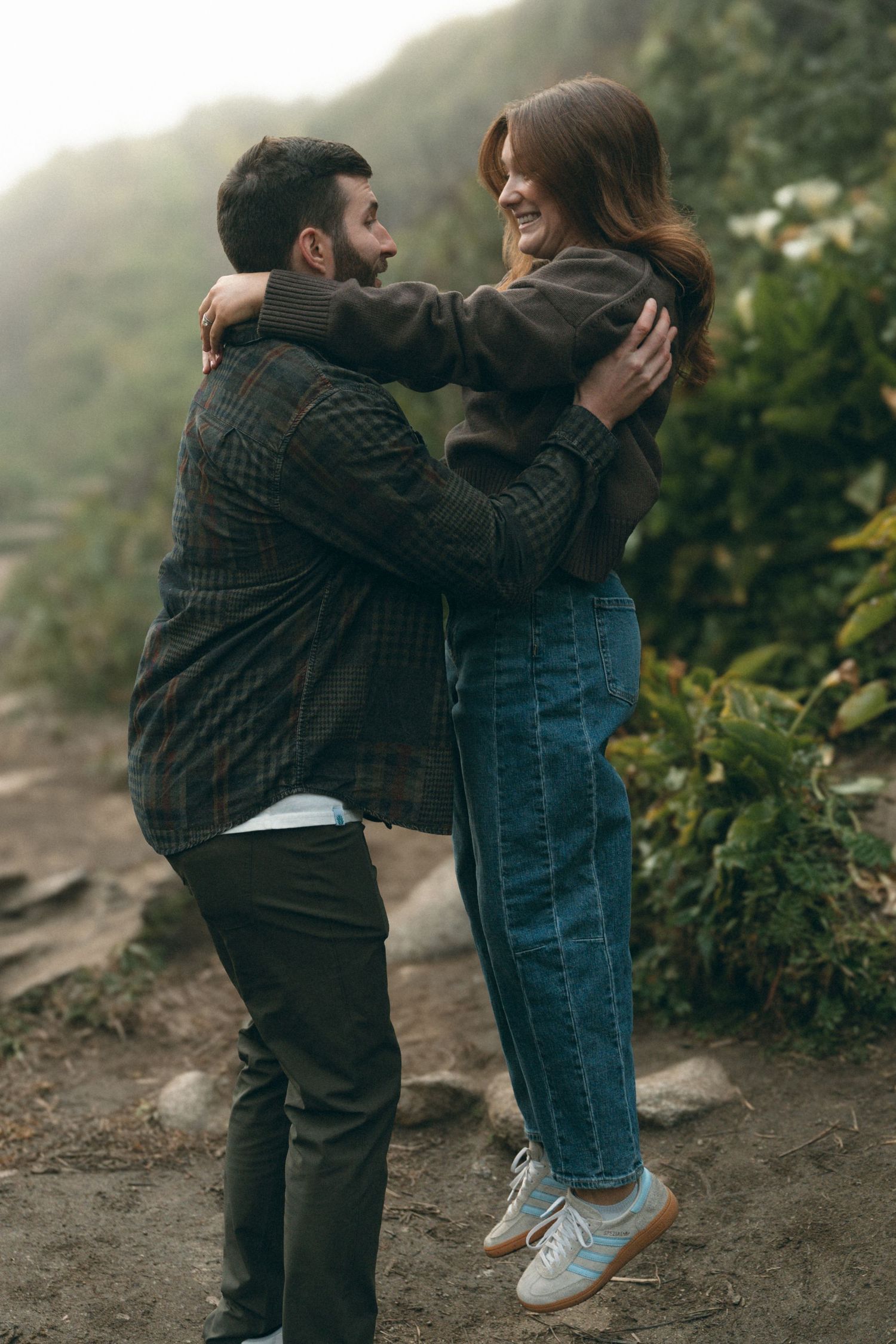 A couple embracing and laughing together on a mountain trail surrounded by foliage.