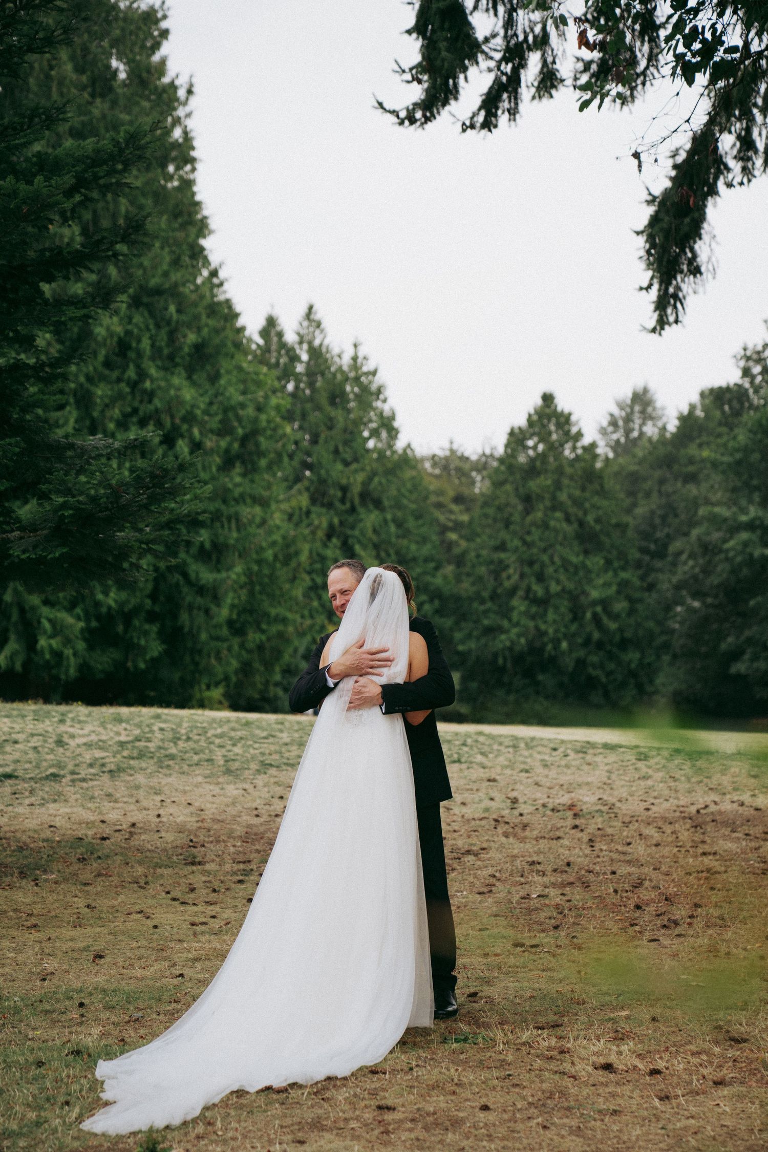 A romantic embrace between newlyweds in white dress and black suit against a backdrop of evergreen trees.