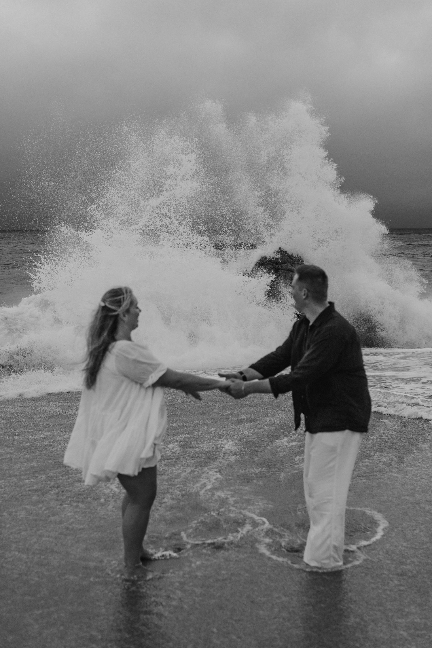 A couple playfully gets caught in ocean waves splashing around them in a sequence of black and white photos.