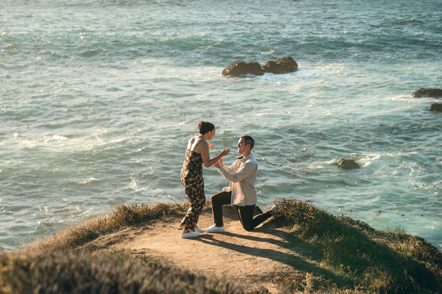 Couple enjoying a romantic moment on rocky coastal cliffs overlooking the ocean at sunset.