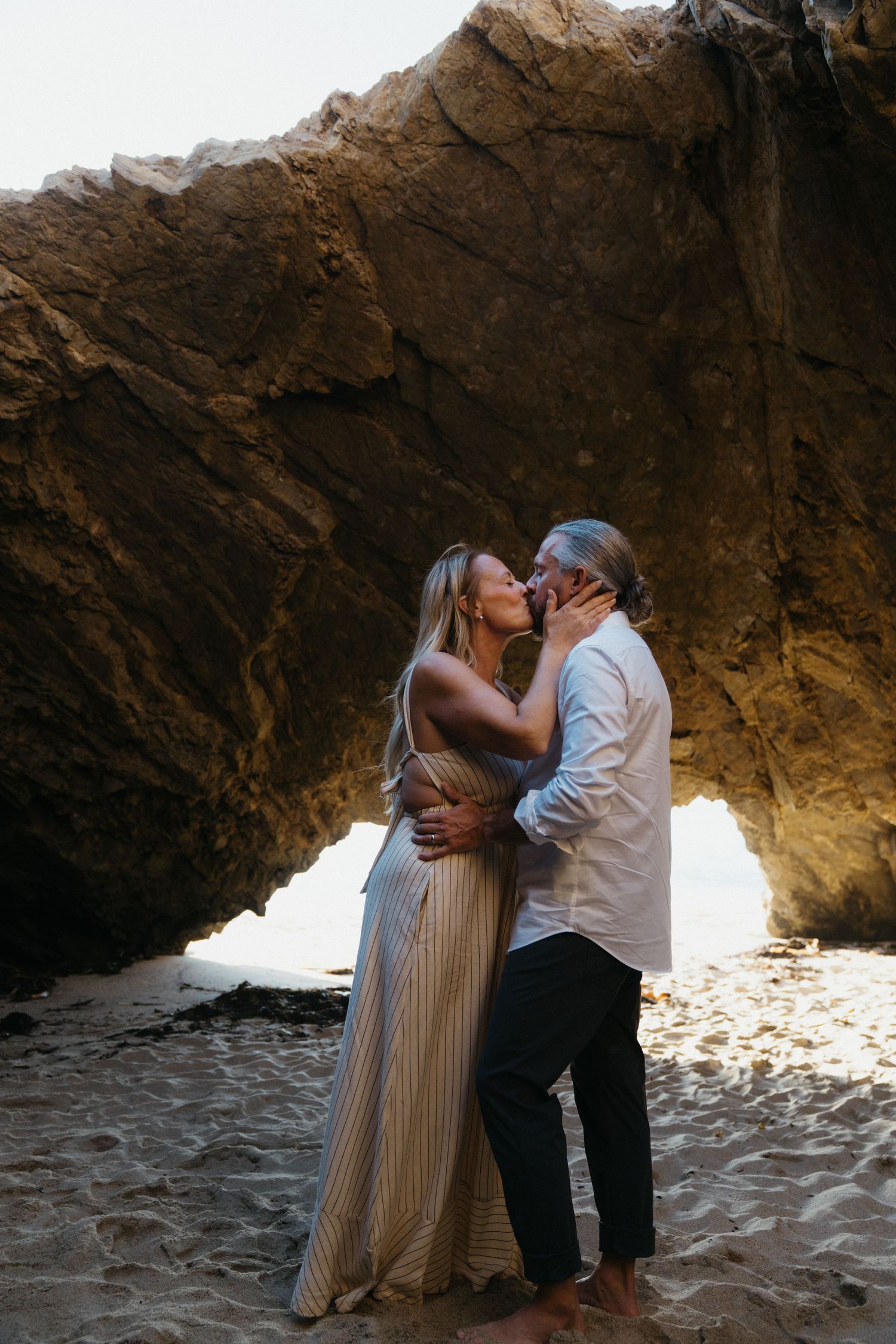 A couple shares an intimate moment while standing in a rocky cave opening with bright sunlight streaming in behind them.