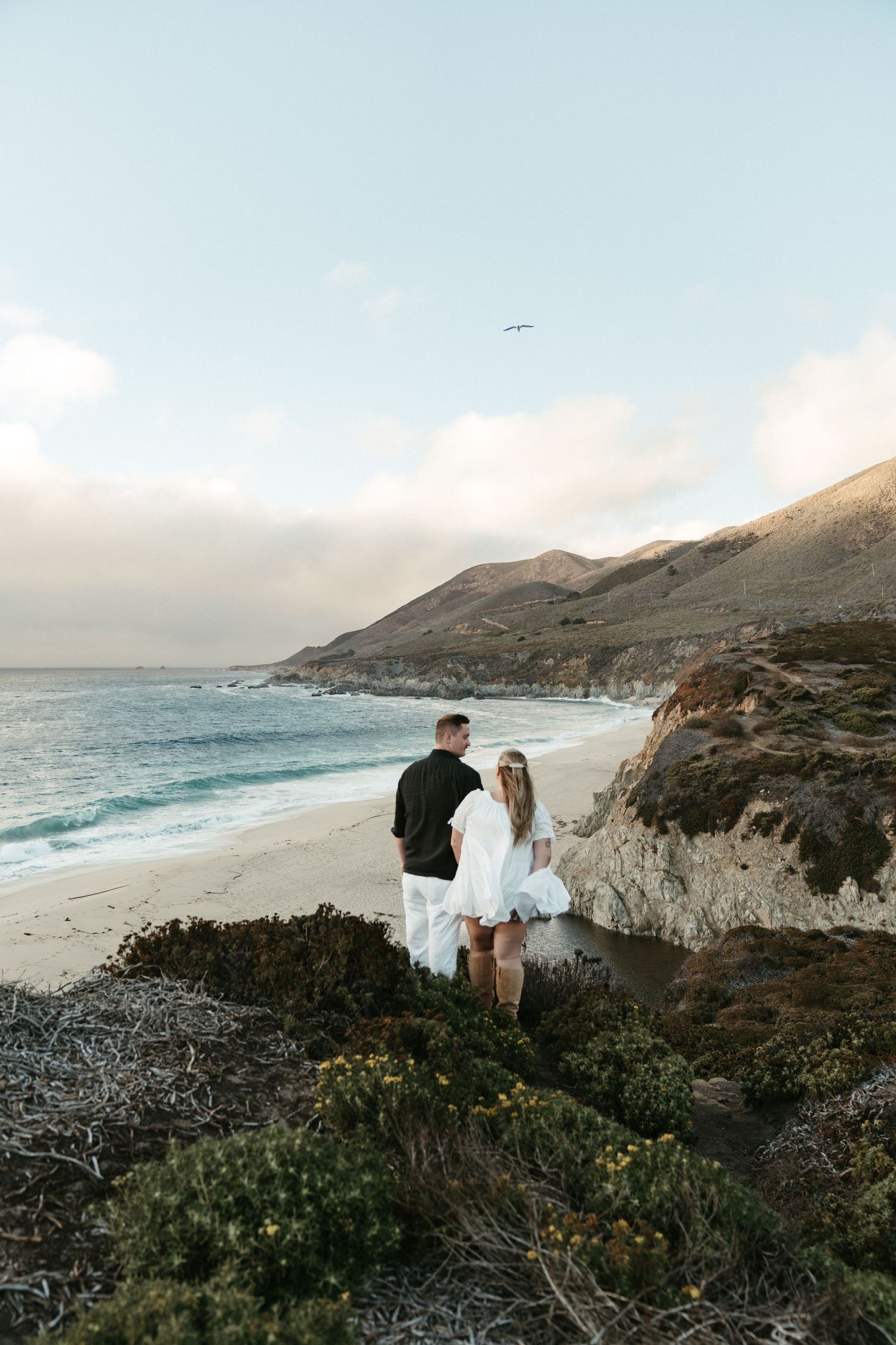 Couple in wedding attire stands on scenic coastal cliffs overlooking Big Sur beach at sunset.