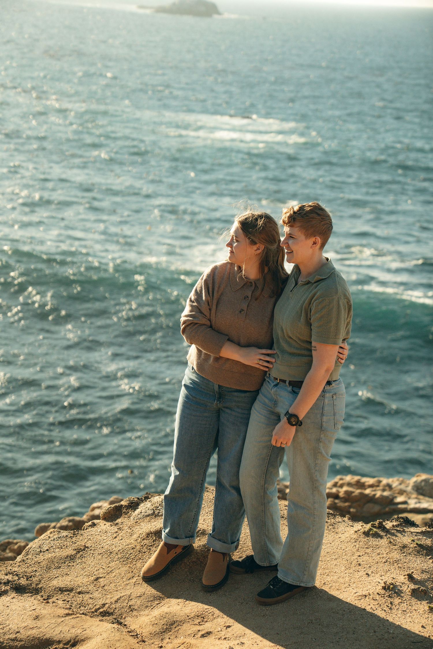 Couple sits close together on coastal cliff edge overlooking the blue ocean waters.