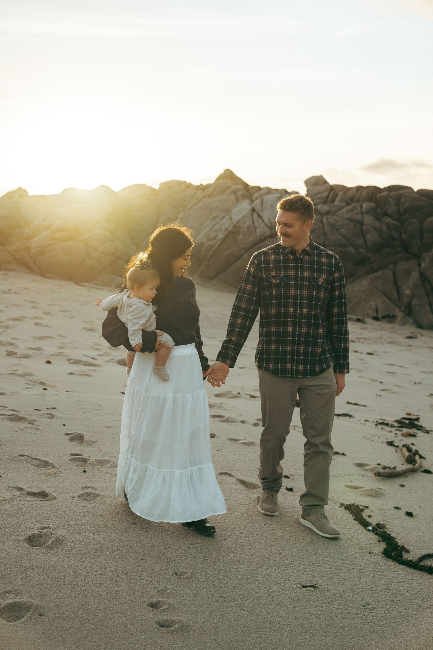 A couple walks hand in hand along the beach shore during golden hour with sunlight behind them.