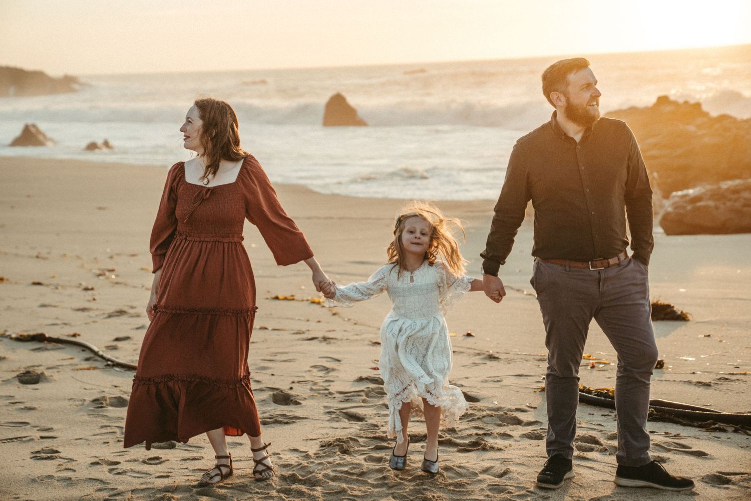A family walks together on a scenic beach during golden hour sunset with waves crashing in the background.