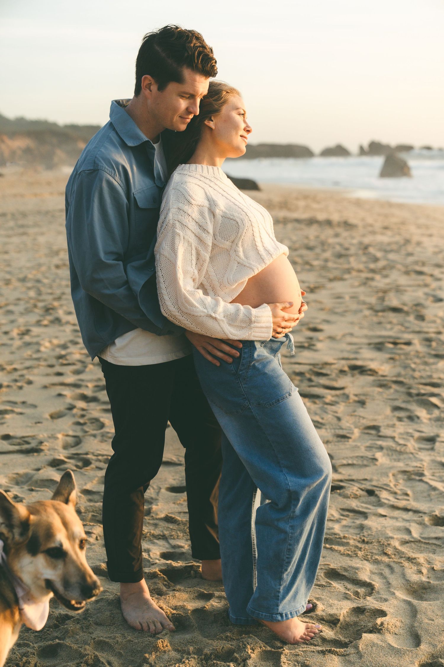 Couple embracing on sandy beach at sunset, pregnant woman in white sweater and jeans, man in denim shirt, dog nearby.