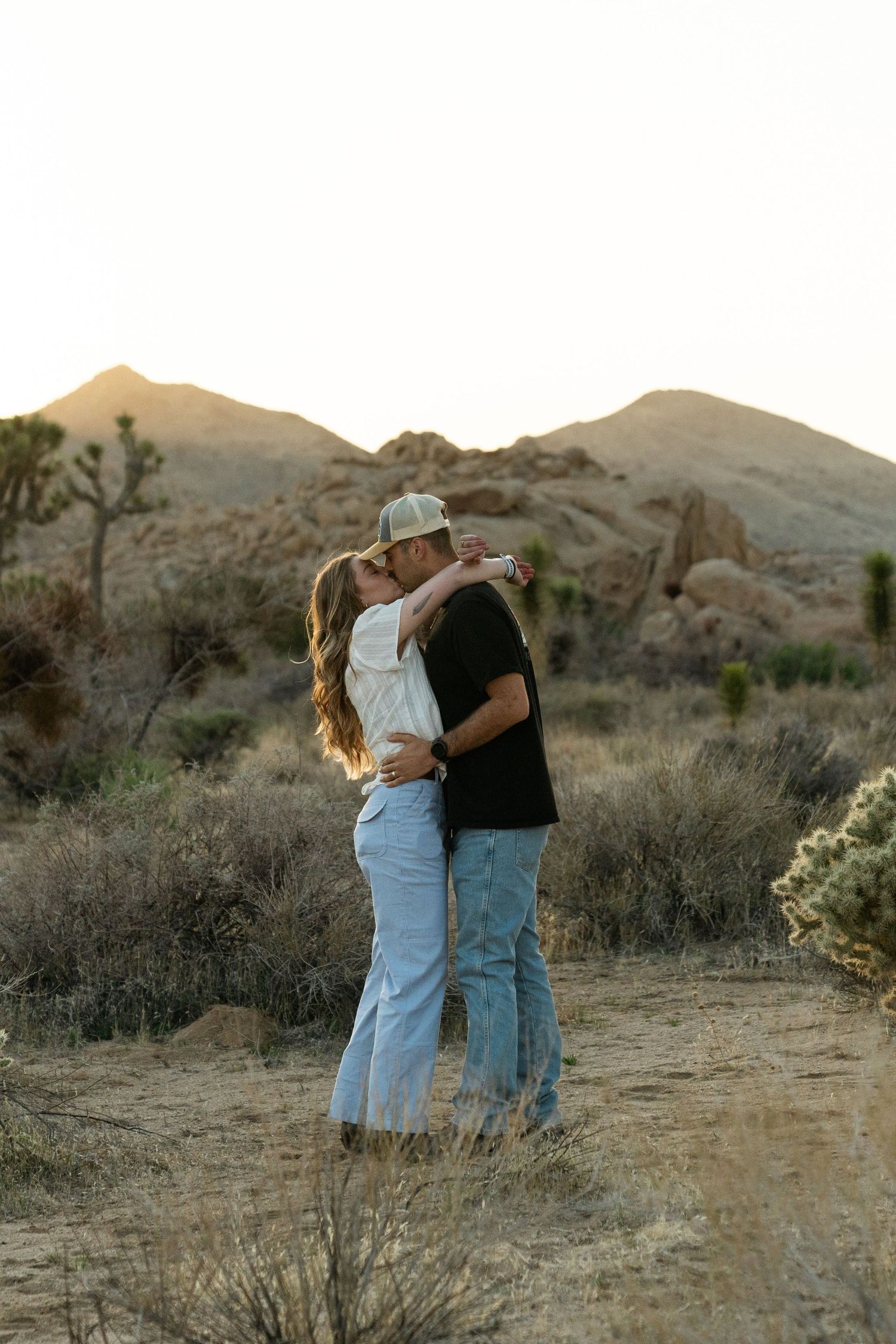 Two people share an intimate moment at sunset in a desert landscape with mountains in the background.