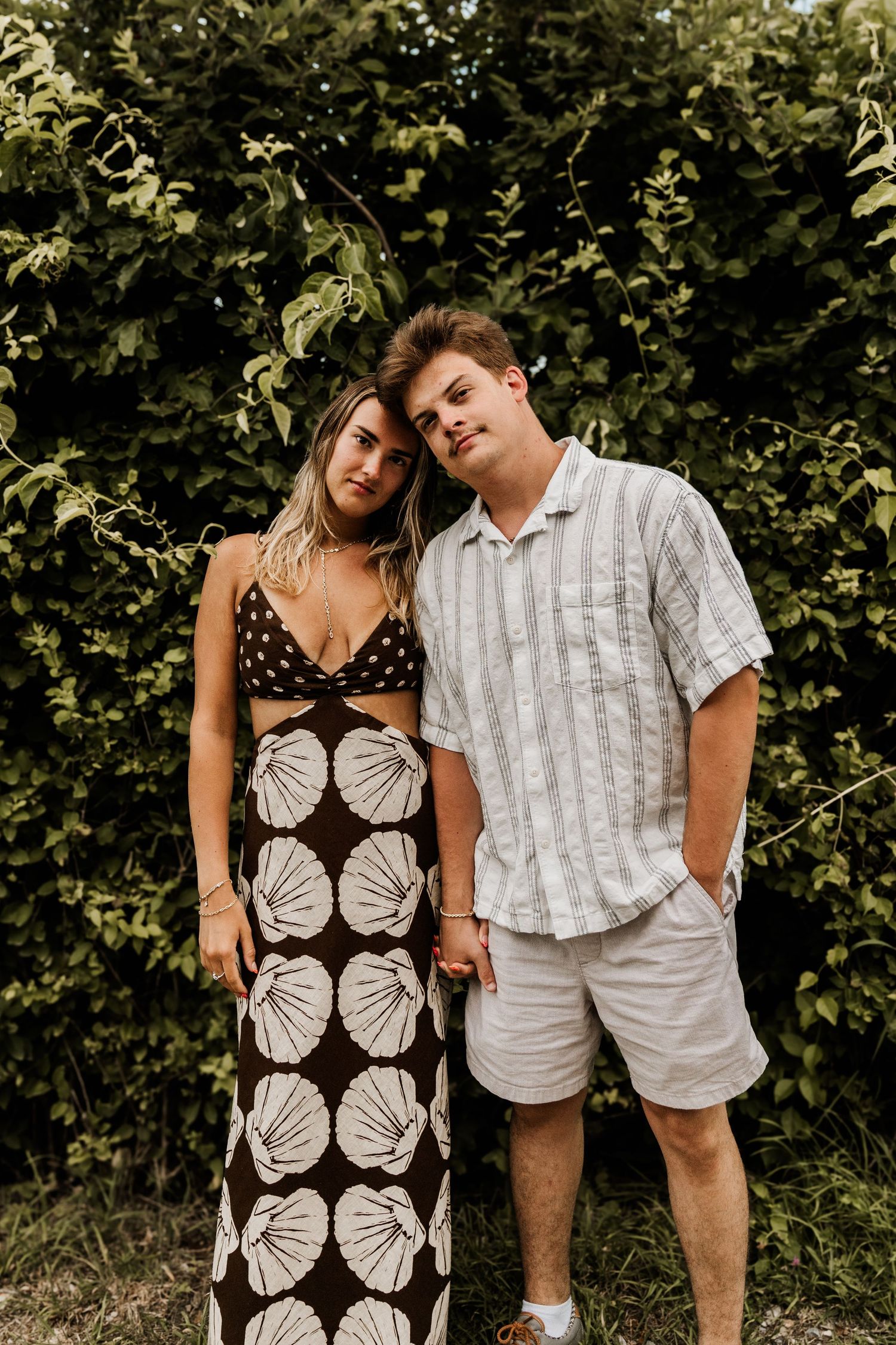 A couple poses outdoors against a leafy green backdrop wearing summer outfits with a black and white patterned dress and white shirt.