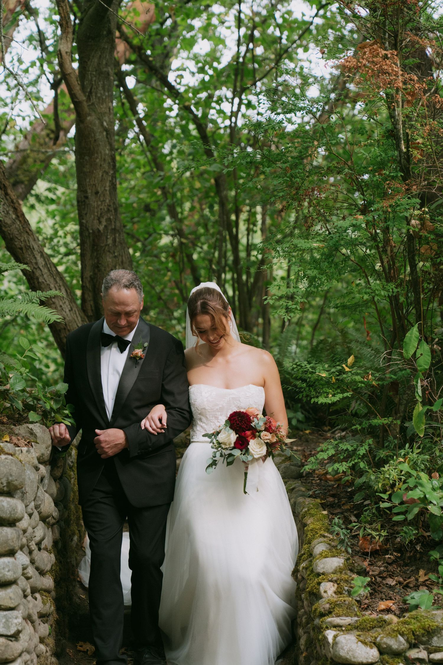 A couple walks together through a wooded path with stone walls during their wedding ceremony.